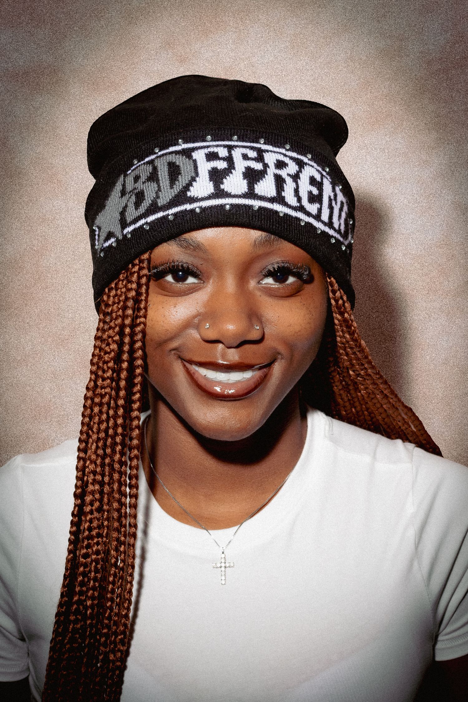 Young woman smiling, wearing a black beanie hat, orange box braids, white t-shirt, and a cross necklace.