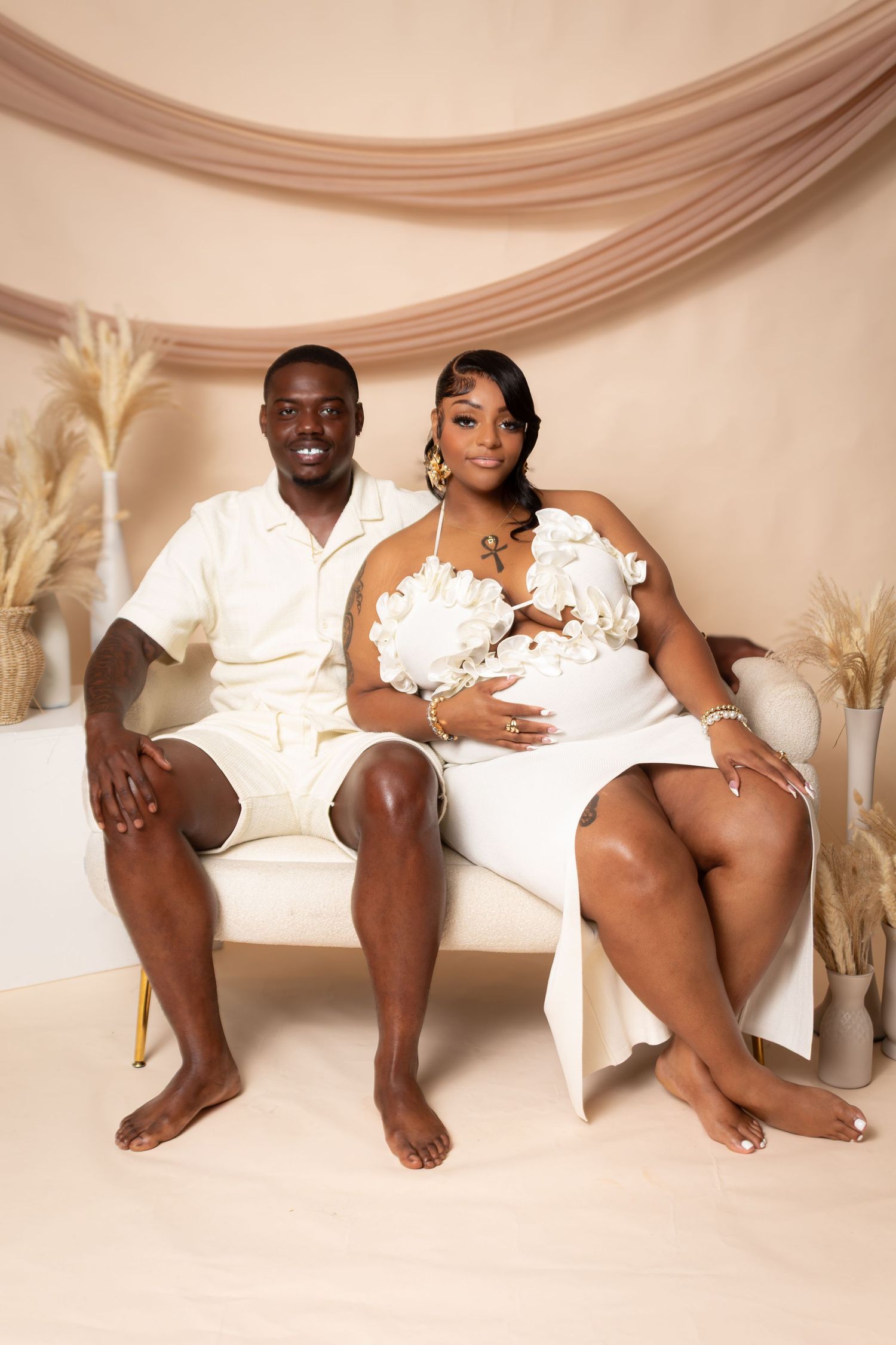 A couple in white outfits poses for a maternity photo shoot in a beige studio setting with dried floral decor.