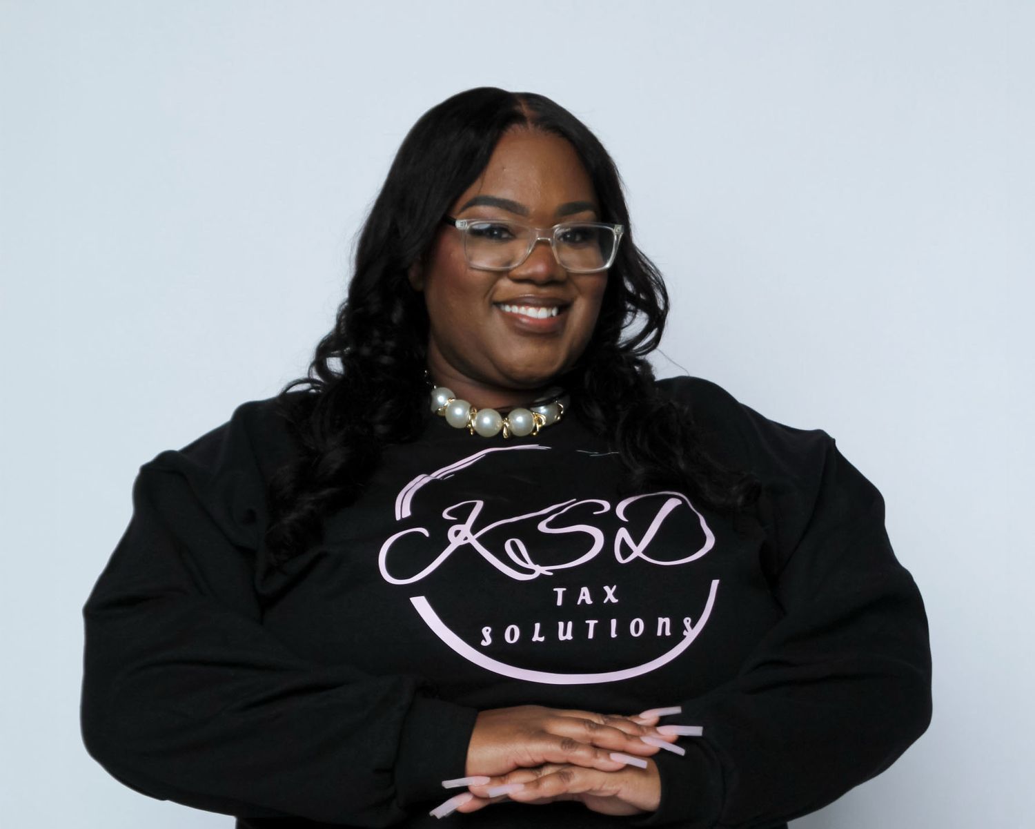 Woman smiling in black KSD Tax Solutions sweatshirt, wearing glasses and pearl necklace, professional photo.