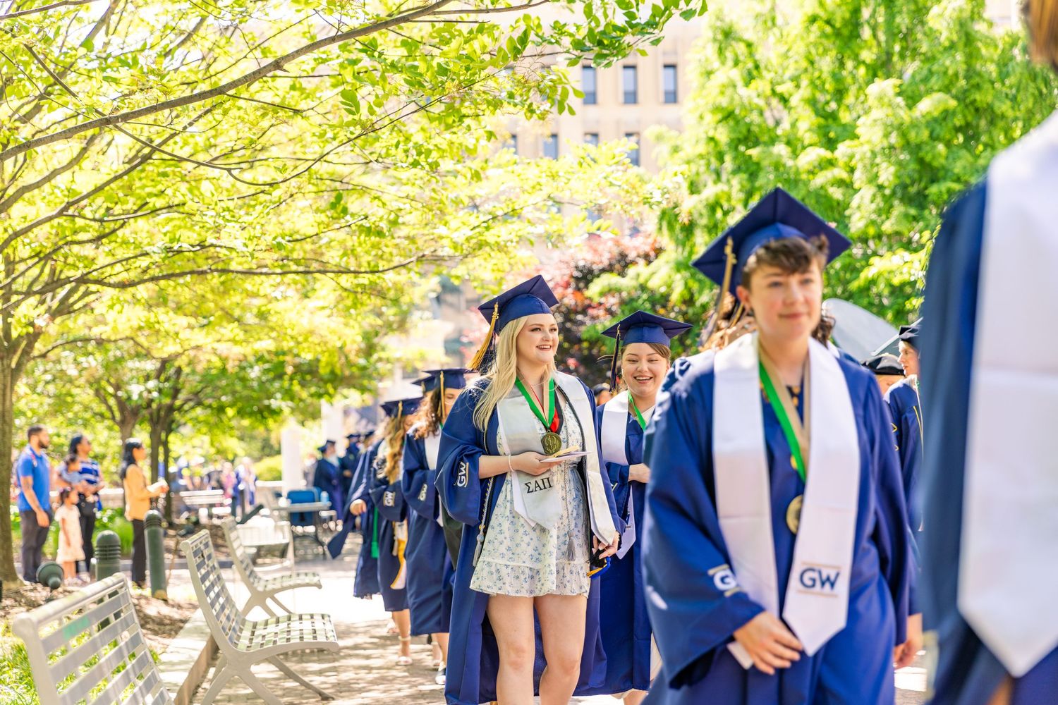 Graduates in blue caps and gowns celebrate during an outdoor commencement ceremony on a sunny spring day.