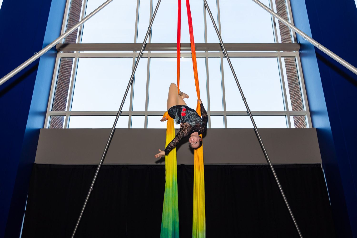 Performer suspended on colorful aerial silks in front of large window.