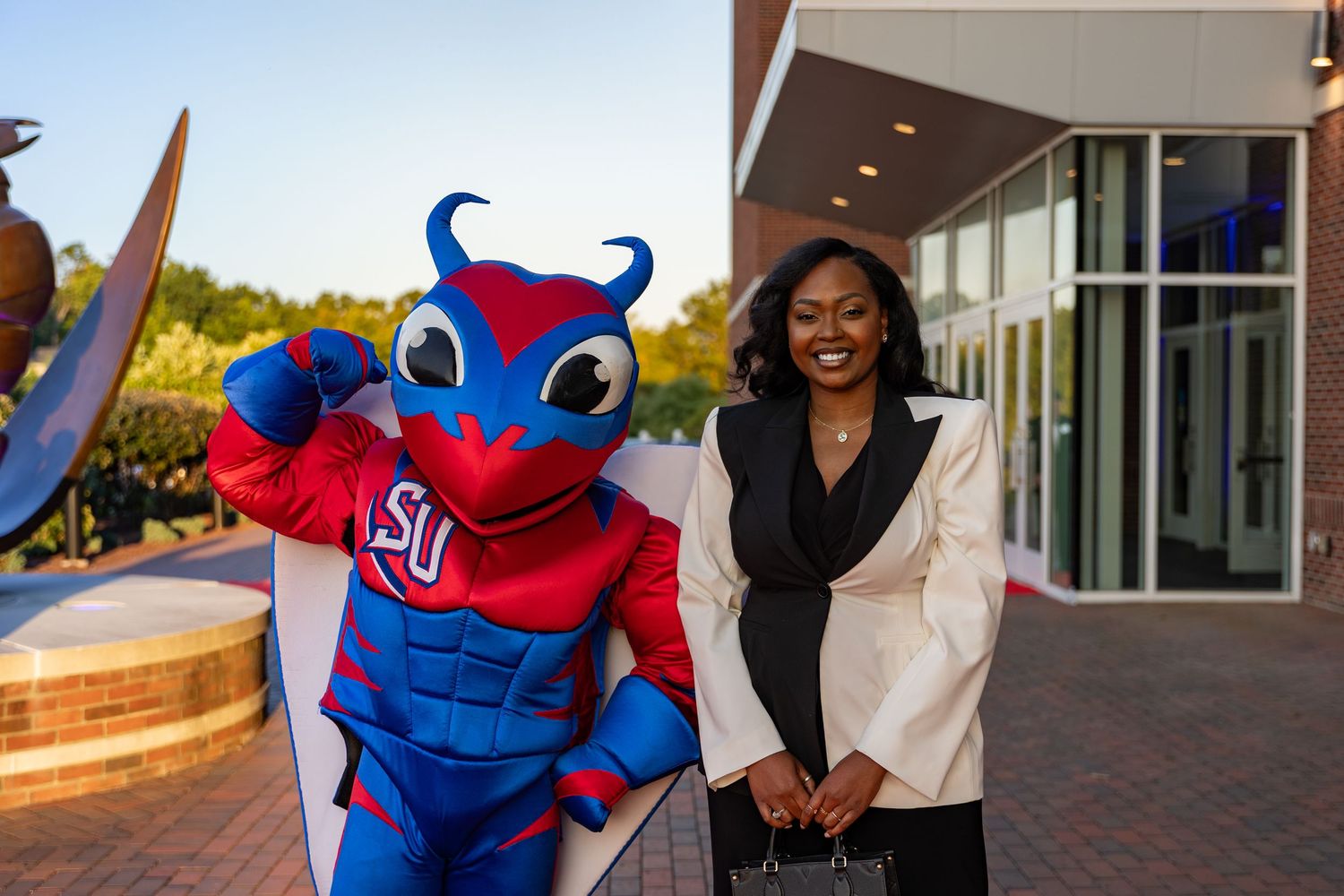 School mascot poses with person outside campus building.