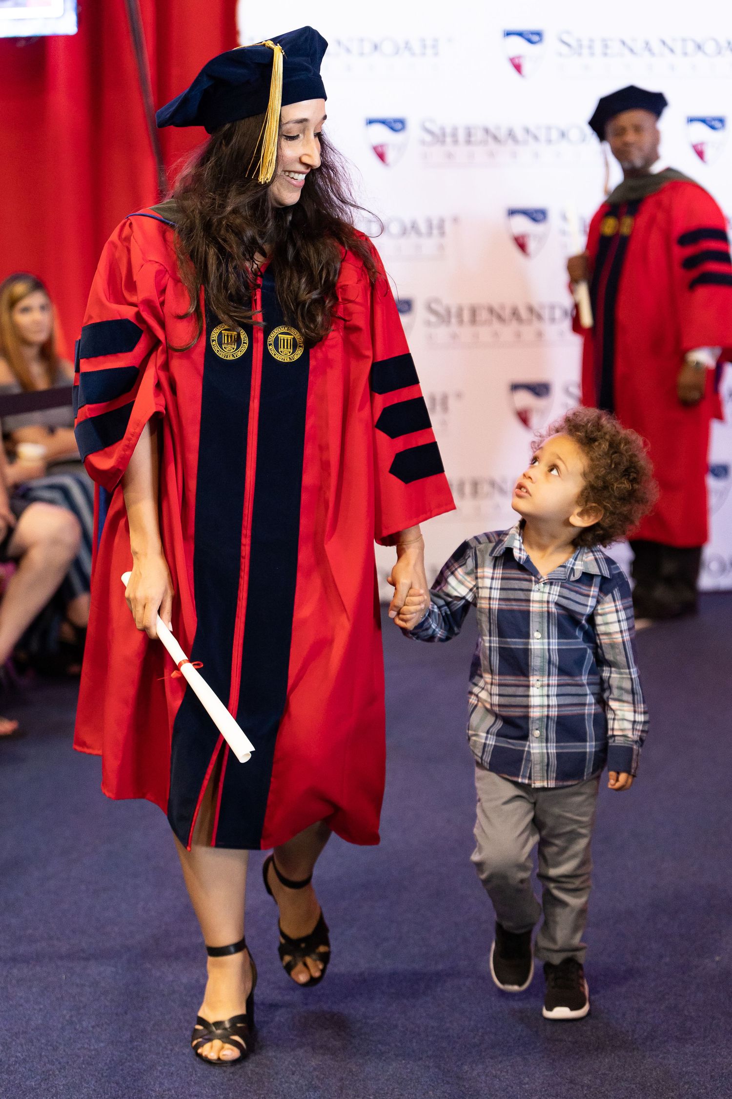 Graduate in red academic regalia walking with small child at commencement ceremony.