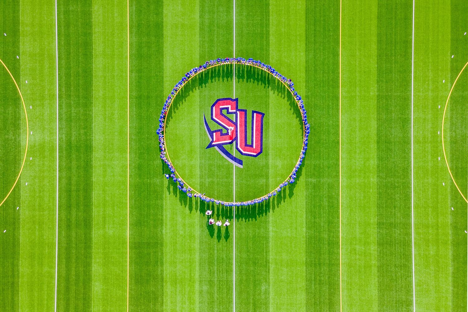 Aerial view of a bright green lacrosse field with members of a marching band gathered in the center.