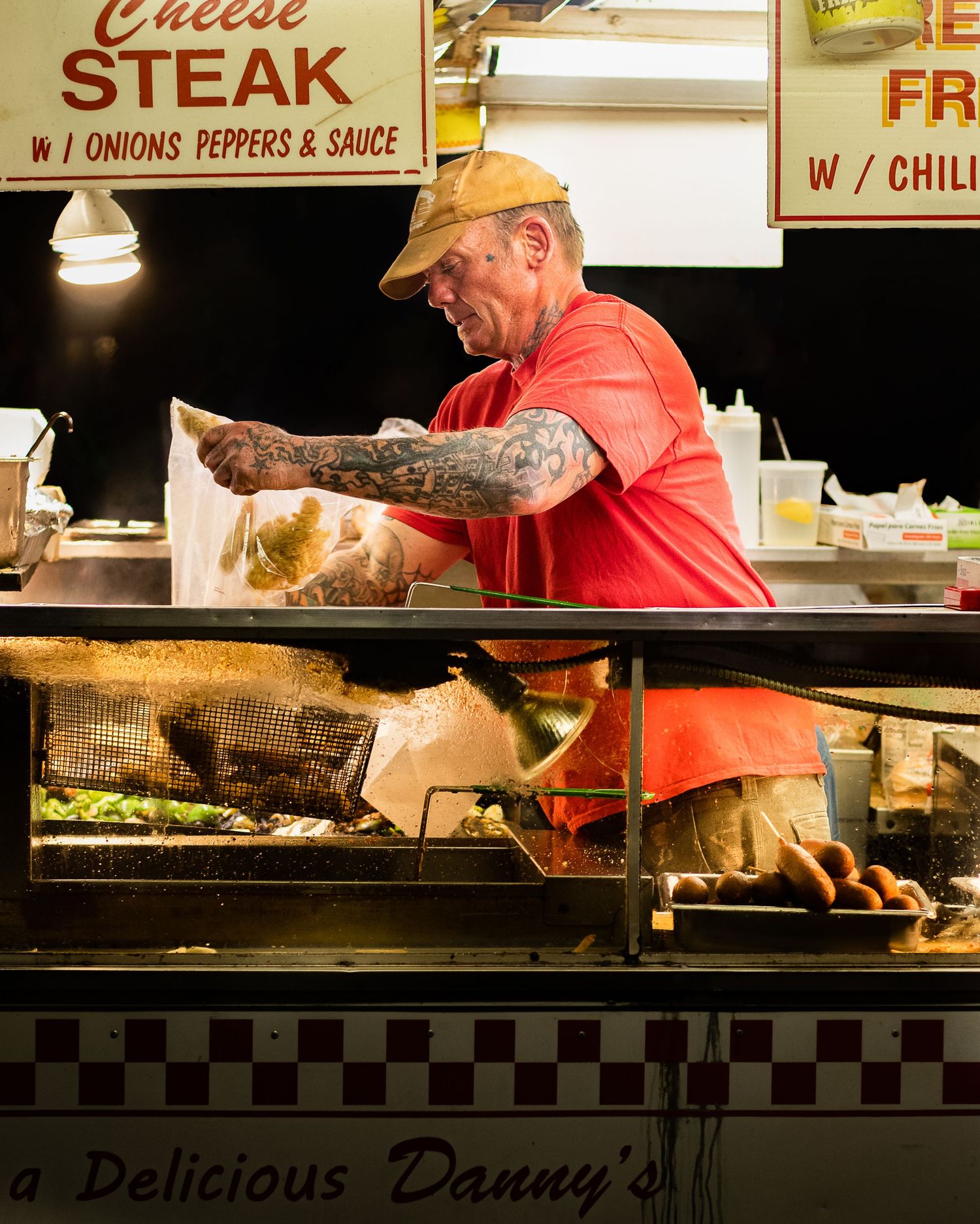 Tattooed cook in red shirt prepares cheese steak with onions and peppers at Delicious Danny's food stand.
