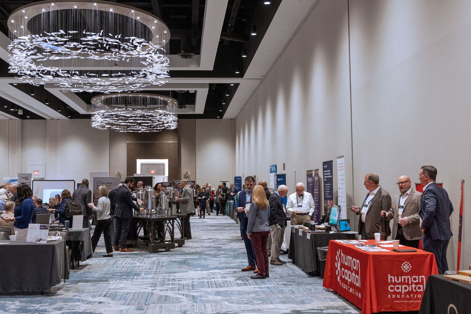 Conference networking event in hotel ballroom with elegant chandelier and vendor tables.