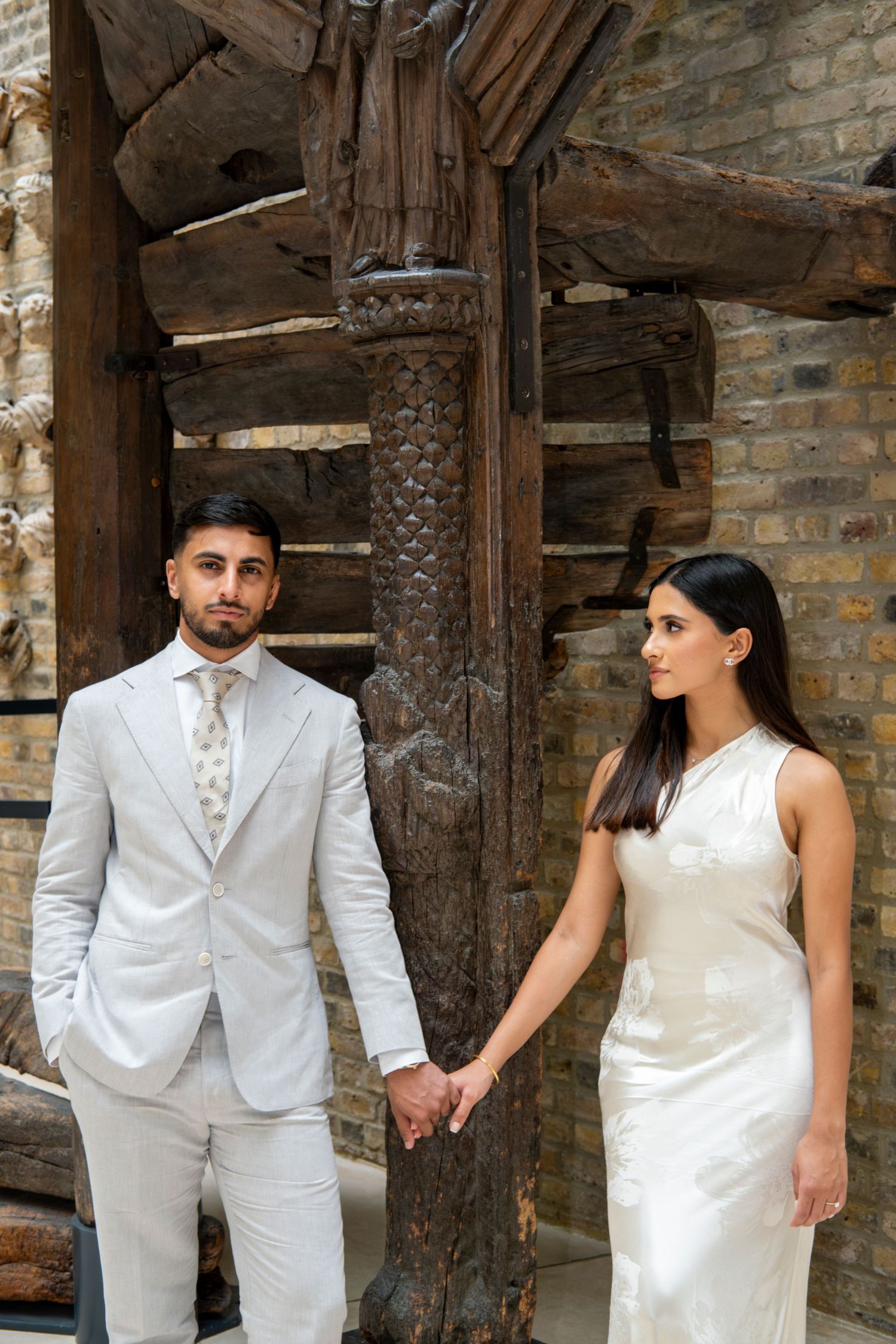 Elegant couple in white holds hands in front of weathered wooden post and brick wall.
