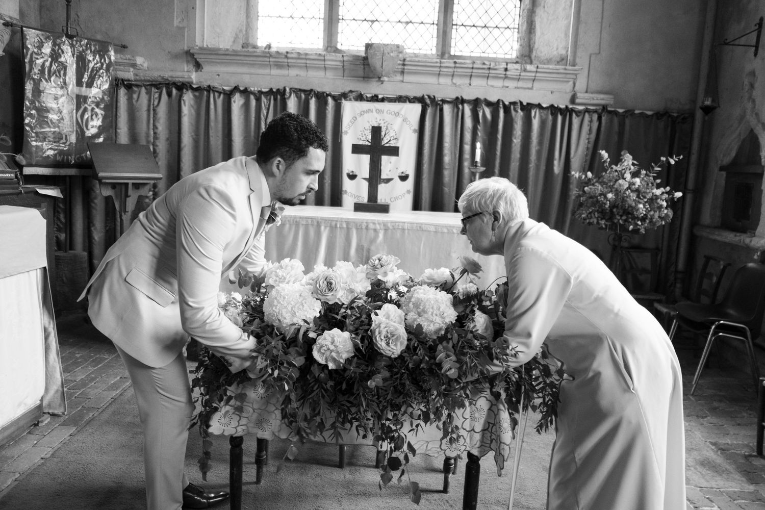 Two people arranging white floral decorations at church altar.