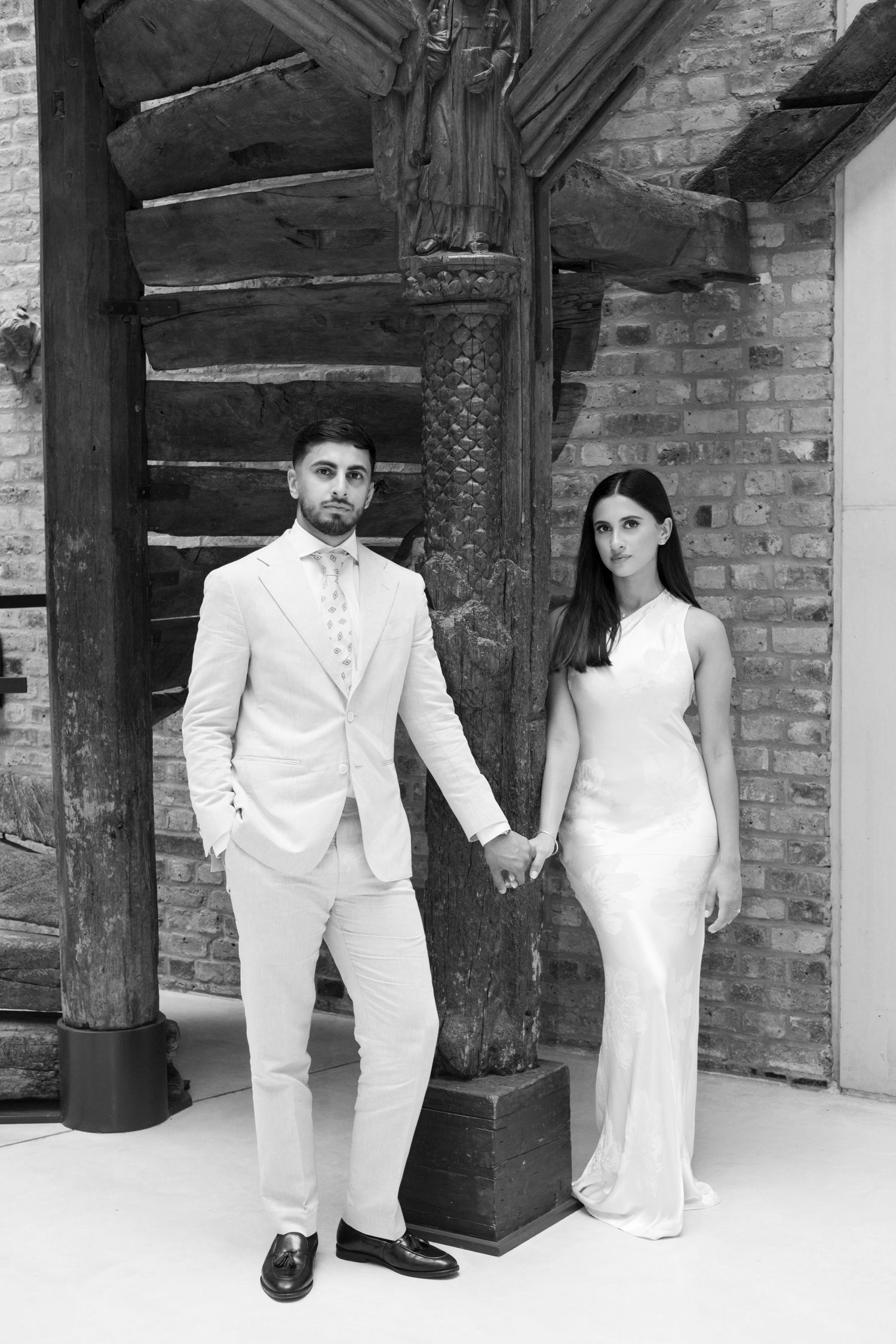 Couple in white formal wear poses in front of rustic wooden beams and brick wall.
