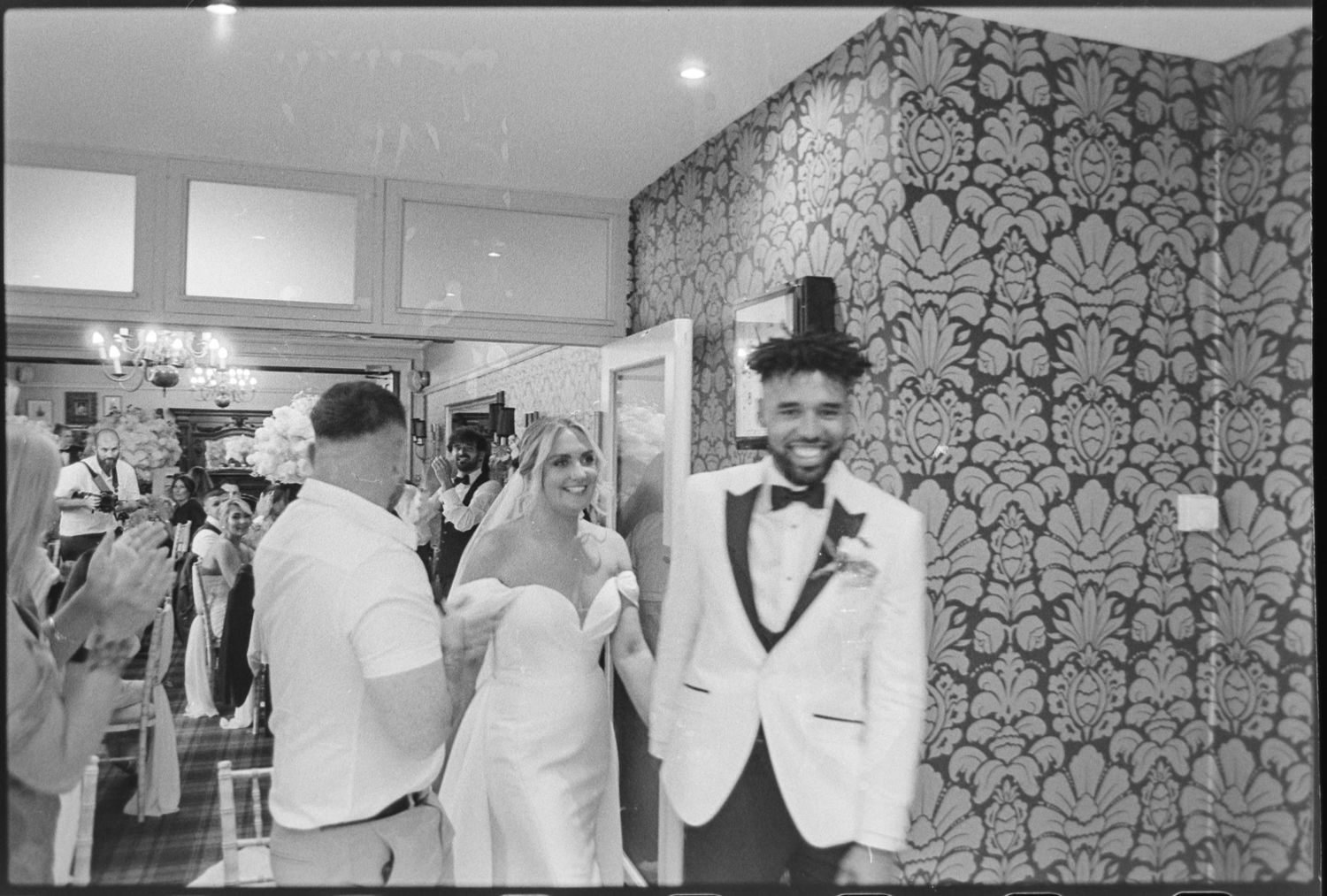 Wedding party members in formal attire gather for black and white photos indoors.