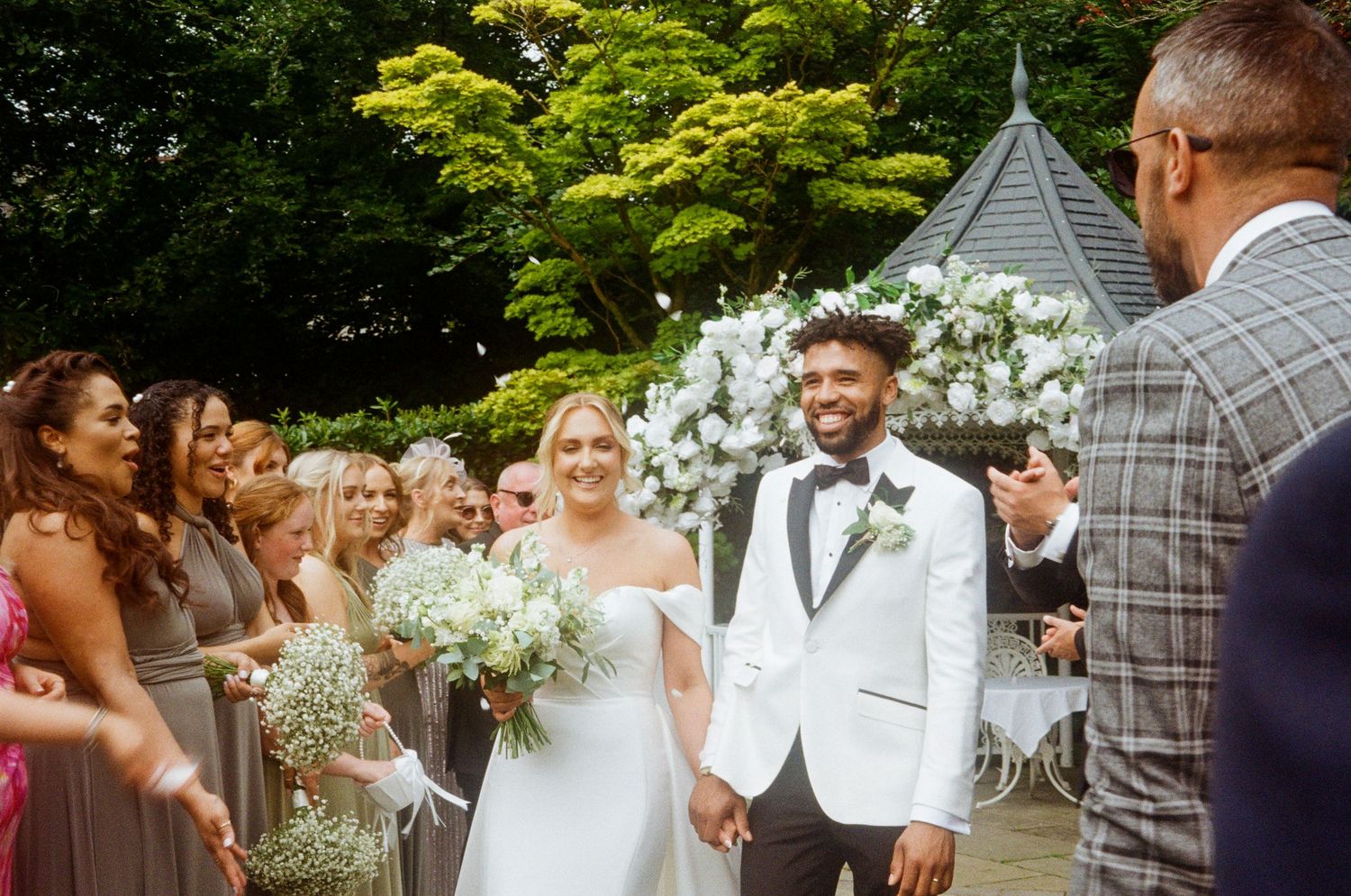 Wedding celebration in a garden setting with guests gathered around decorative floral arrangements.