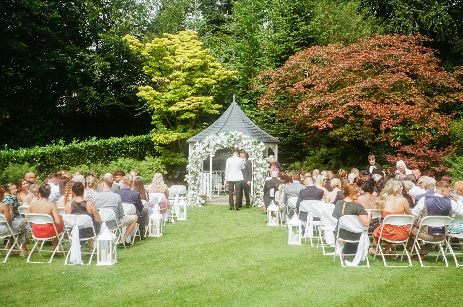 Outdoor wedding ceremony setup with white chairs and a charming gazebo surrounded by lush green trees.