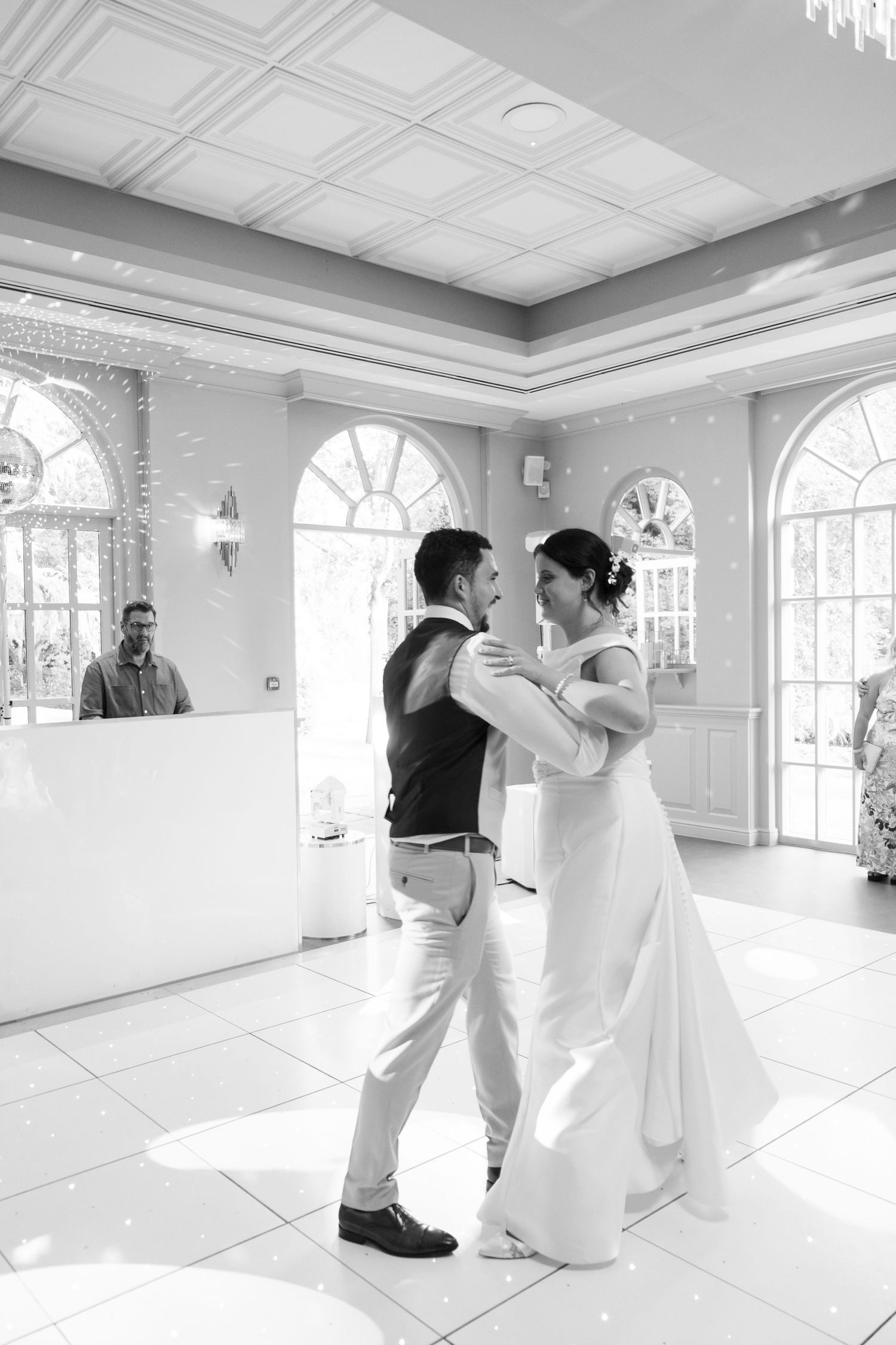 Couple dancing in an elegant white ballroom with arched windows and ornate ceiling details.