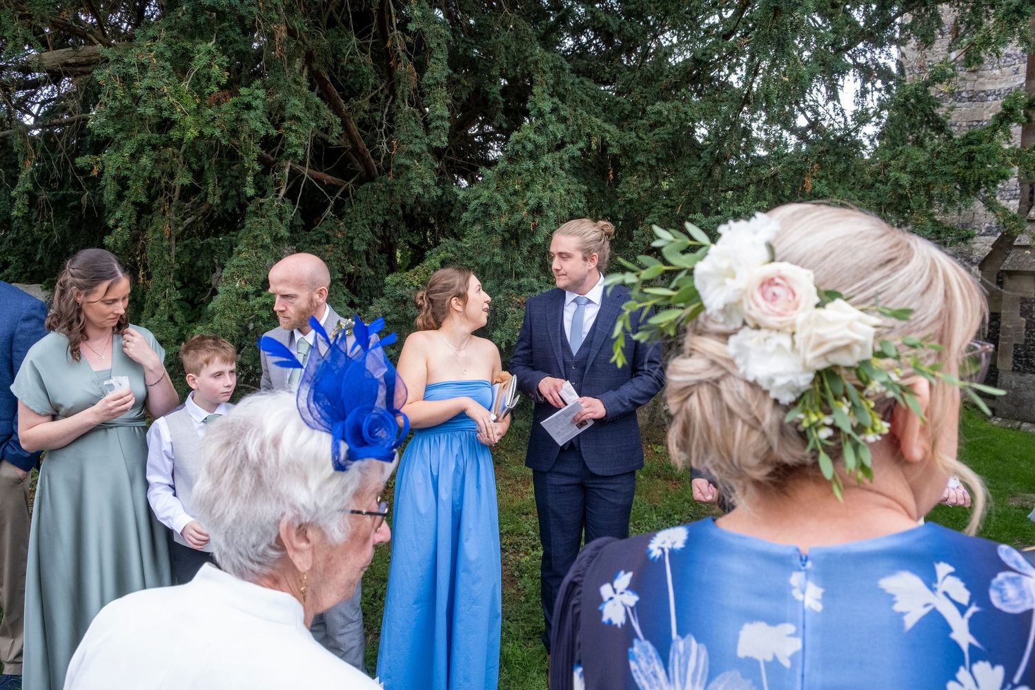 Wedding guests in colorful attire gathered outdoors among trees and greenery.