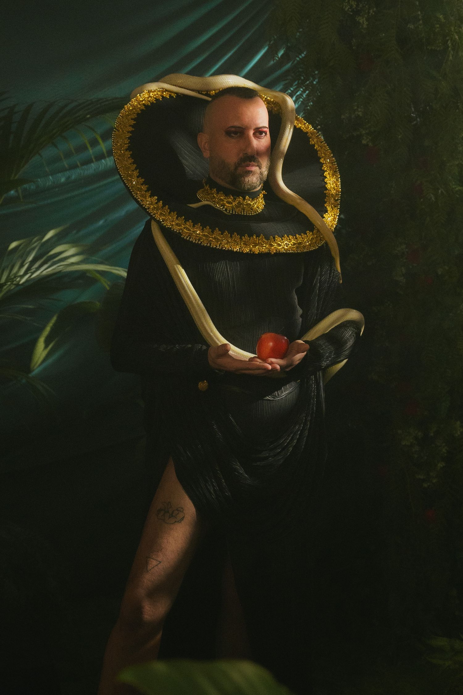 Figure in dramatic Renaissance-style costume with ruffled collar holds red object against dark foliage background.
