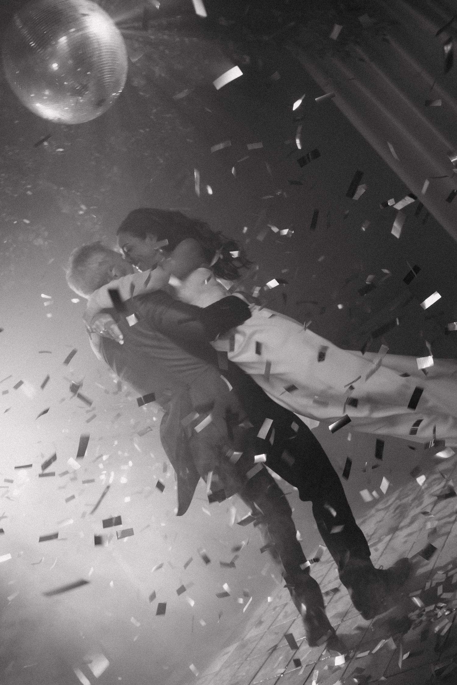 A couple embraces on a dance floor with confetti falling and a disco ball glowing in dramatic black and white lighting.