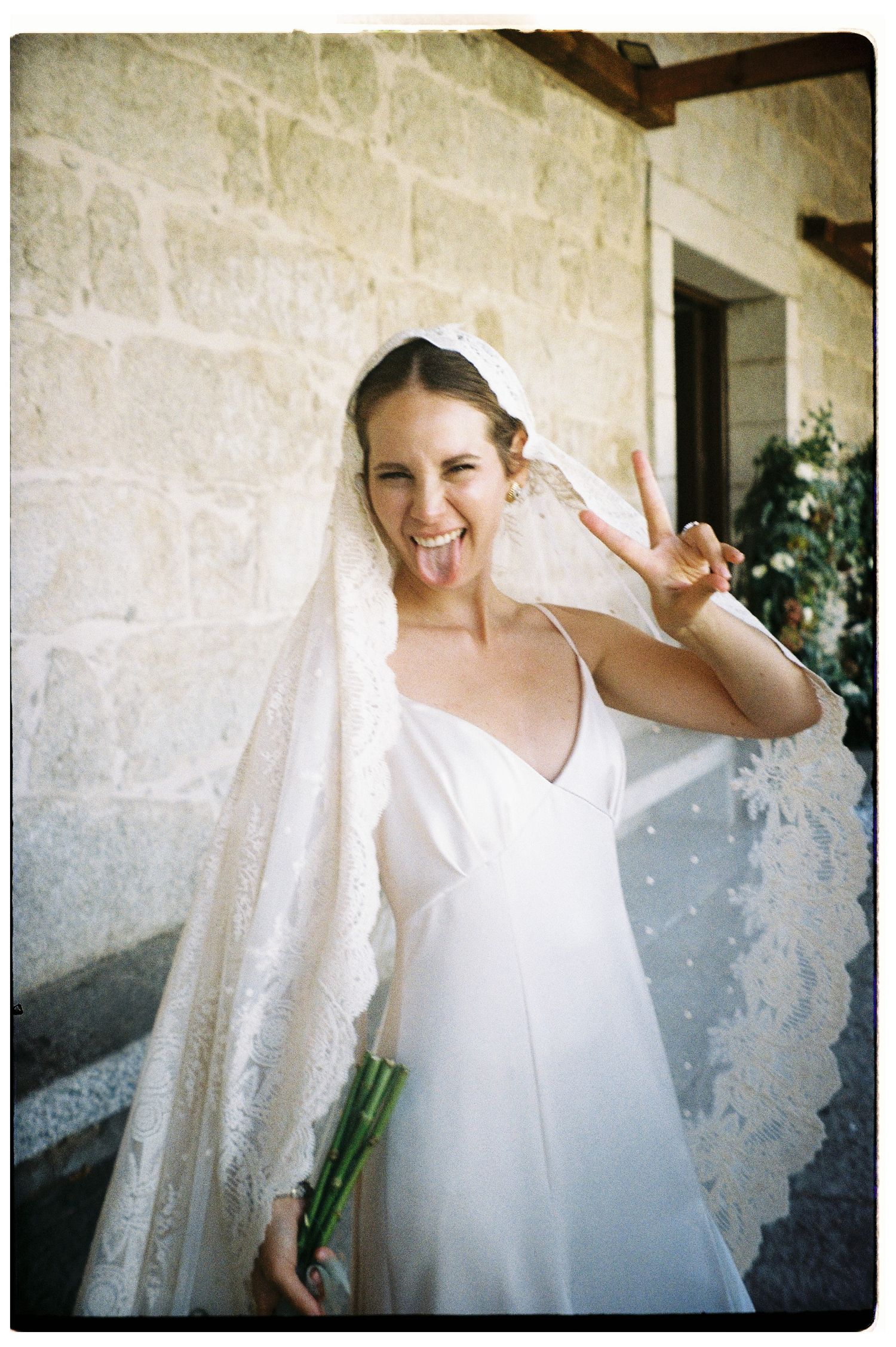 A bride in a simple white dress poses against a textured stone wall with natural light.