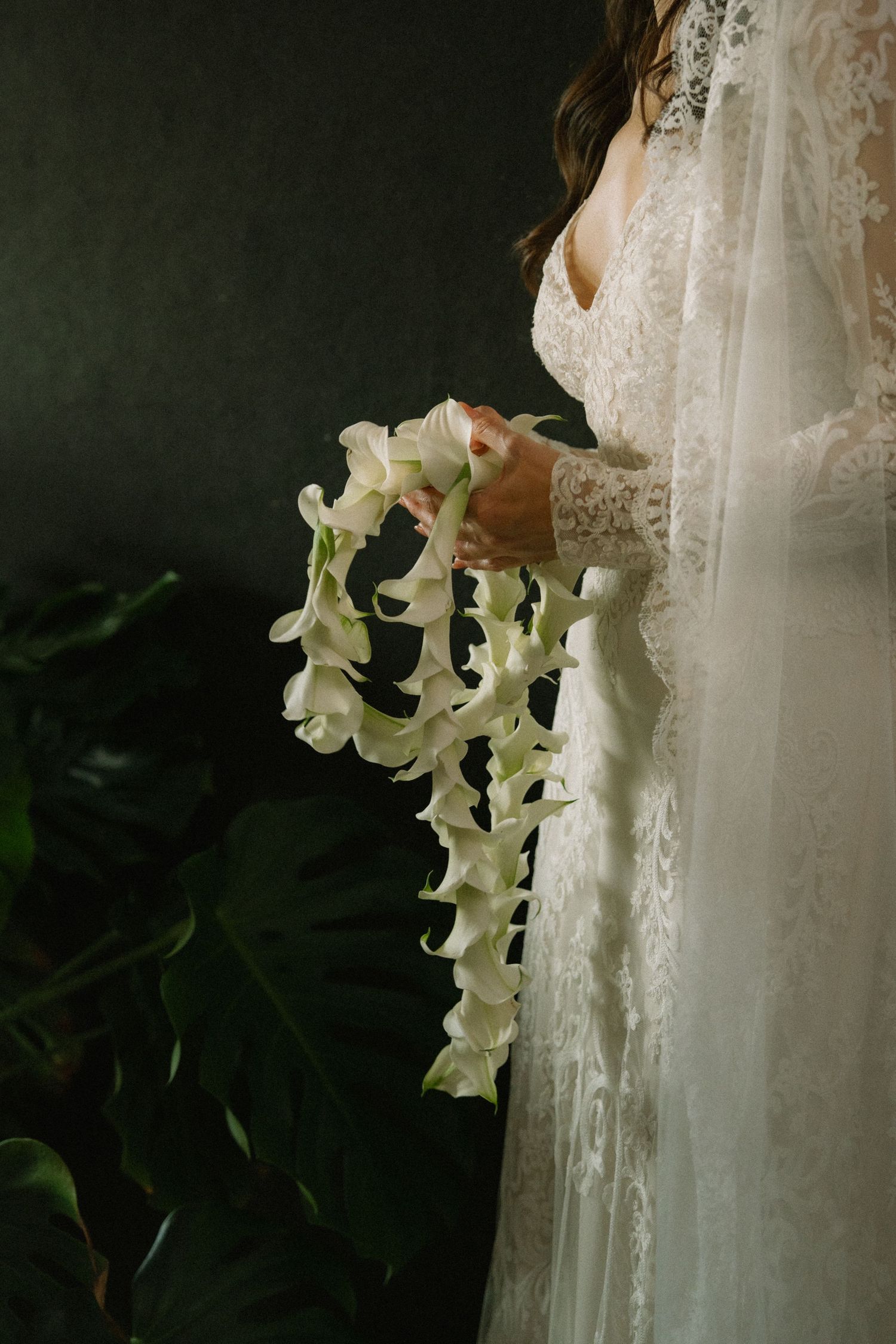 A white wedding dress with delicate lace details is adorned with cascading white lilies against a dark background.