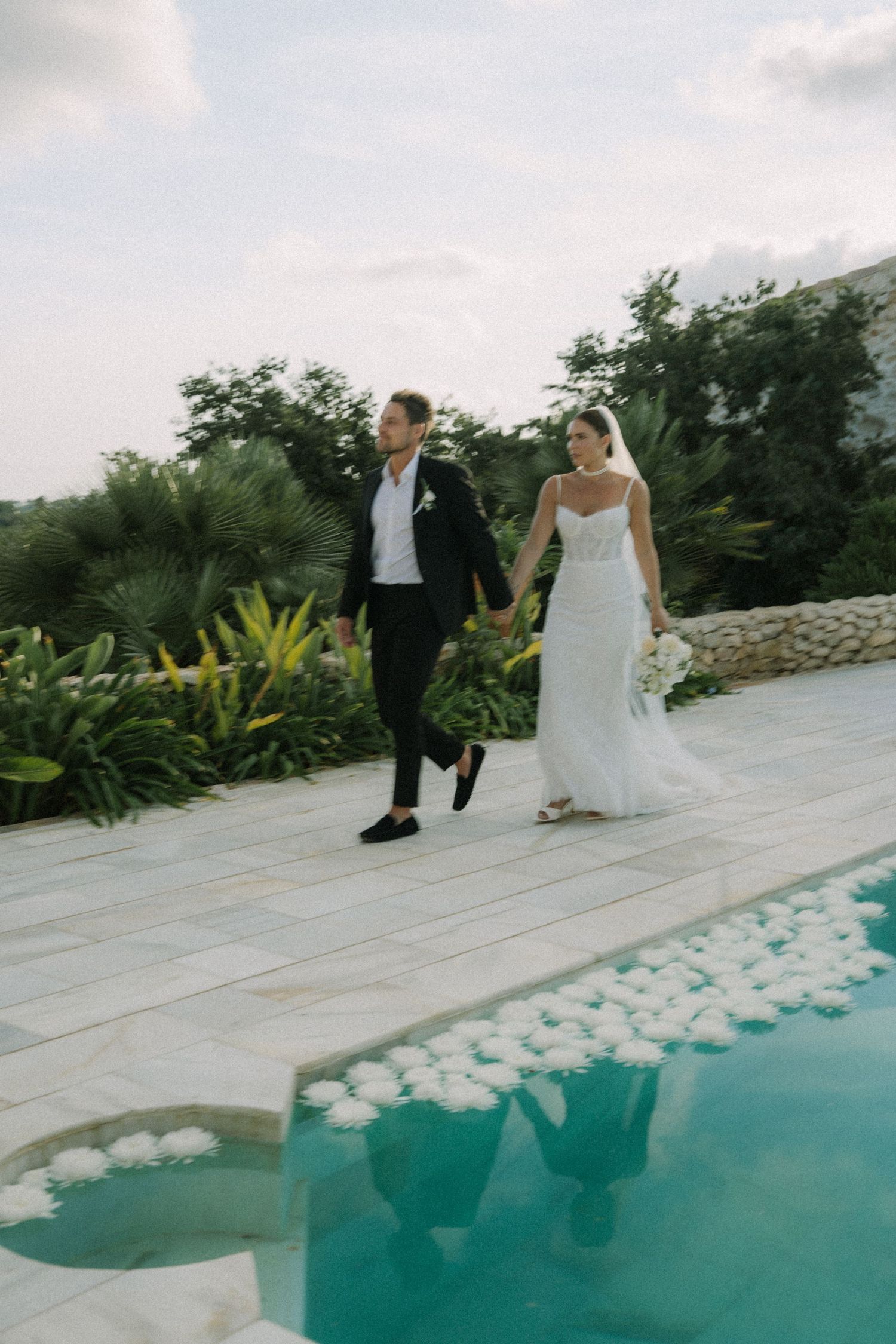 Newlyweds walking along poolside with white flower petals scattered on deck.