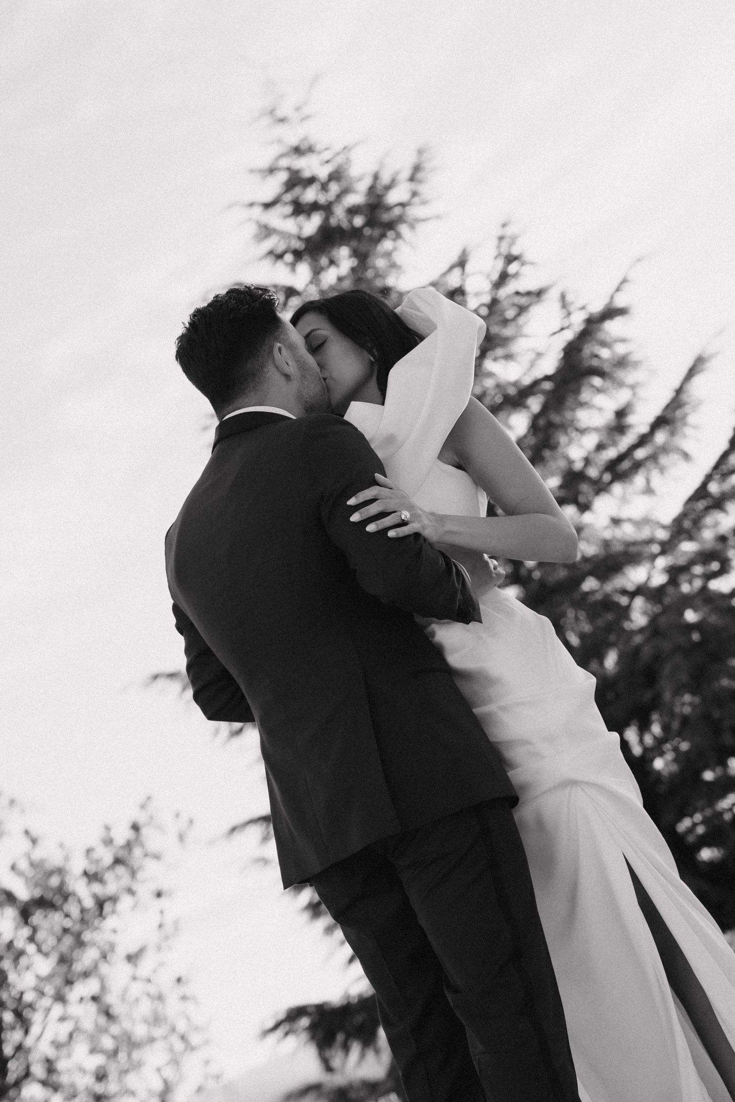 Black and white wedding photos show a romantic kiss between newlyweds with evergreen trees in the background.