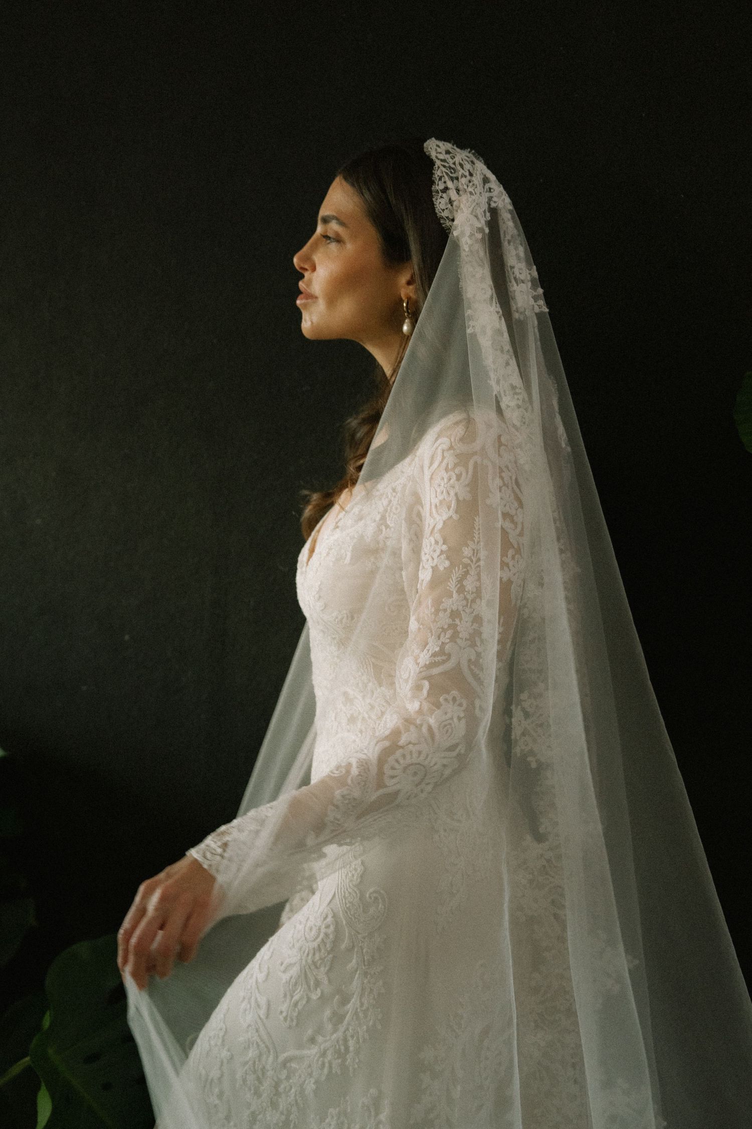 Side profile views of bride in lace wedding dress and flowing veil against dark background.