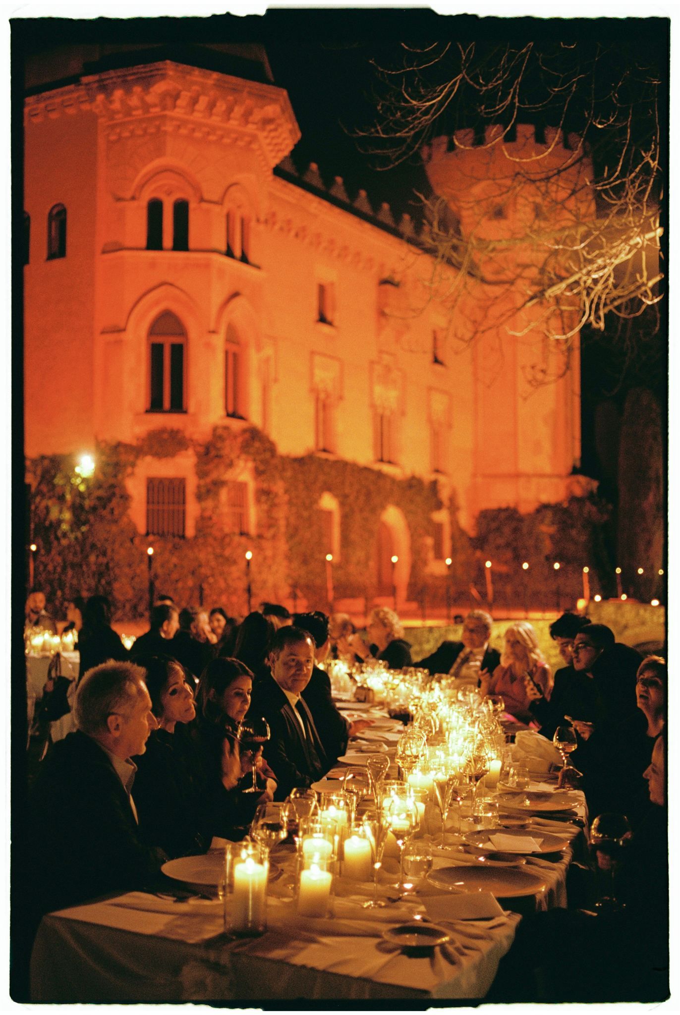 Elegant candlelit outdoor dinner party at dusk with long tables set against a historic building facade.