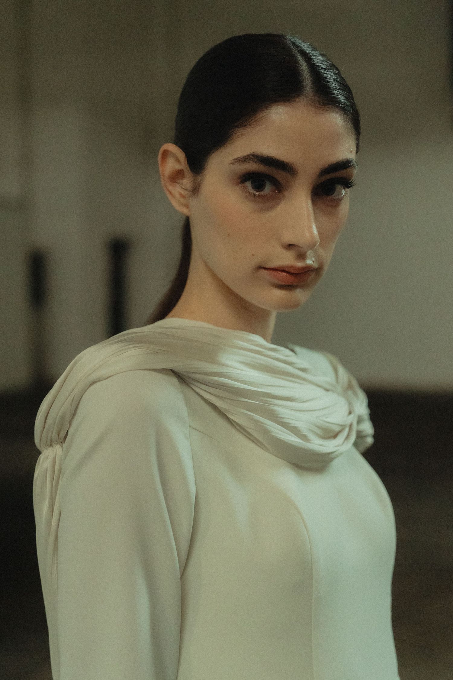 Woman in elegant white dress with draped neckline poses in moody lighting.
