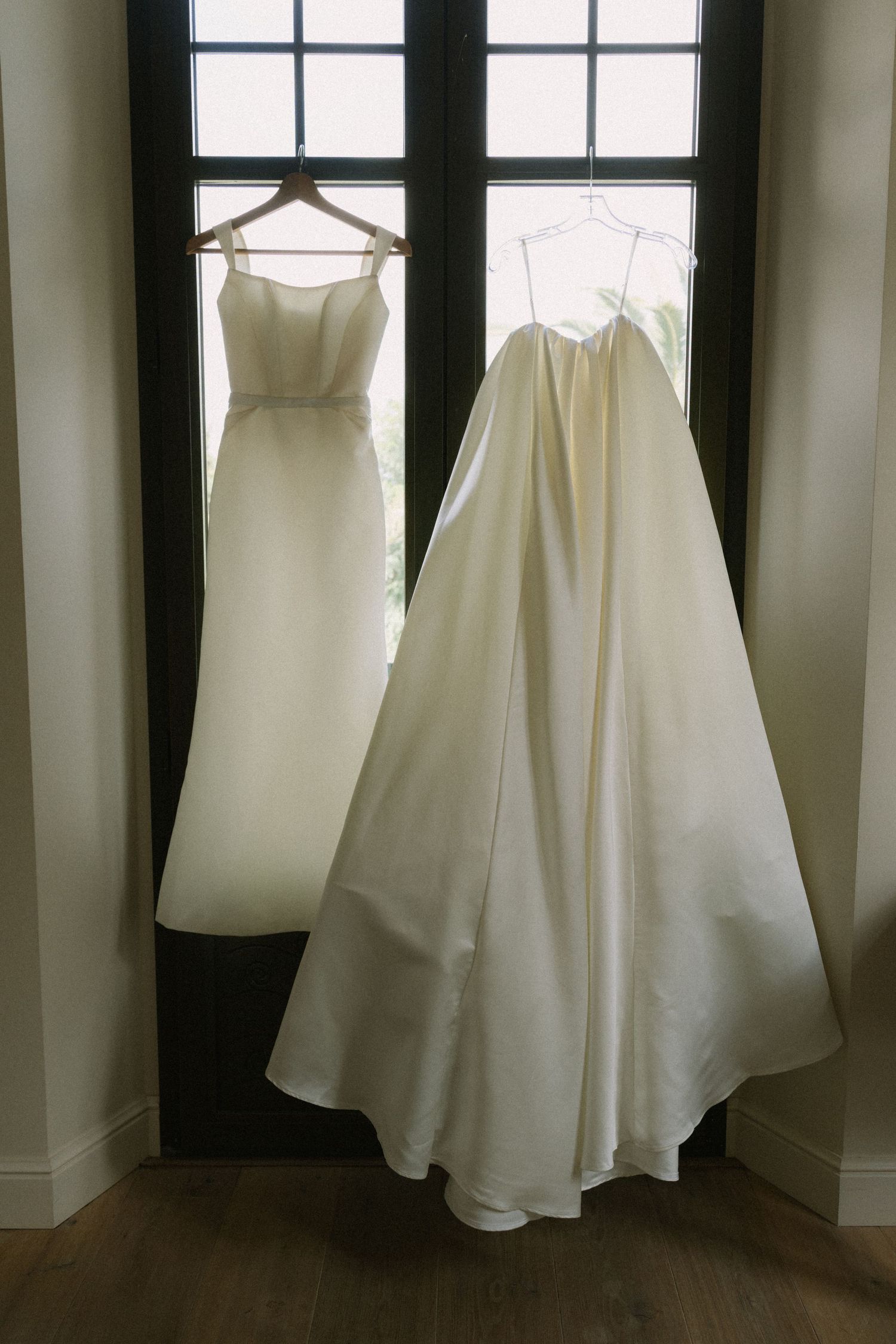 Multiple white wedding dresses and gowns hanging in a row against a dark wall with windows.