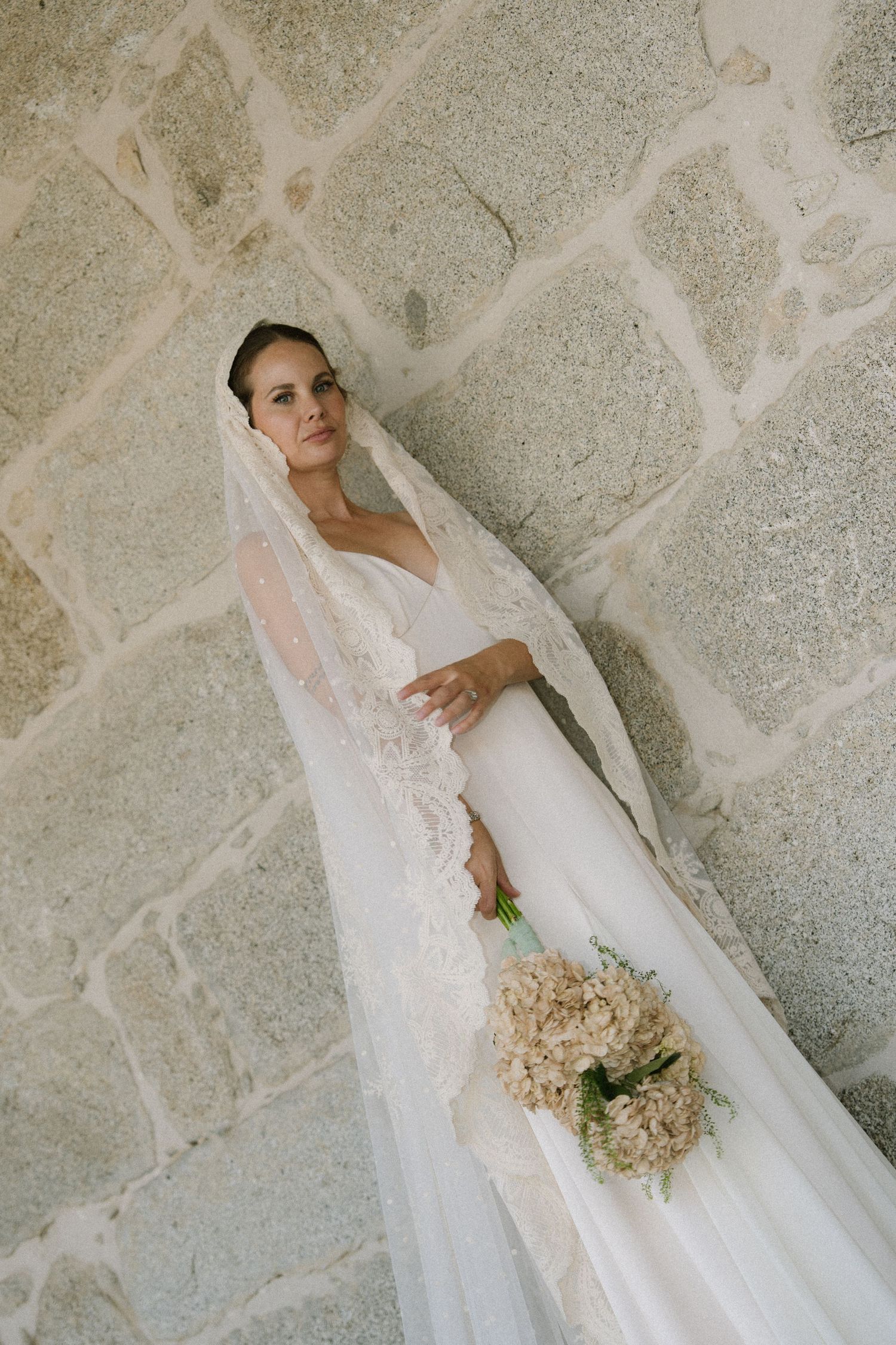 A bride in an elegant white wedding dress and veil poses against a stone wall while holding a pale bouquet.