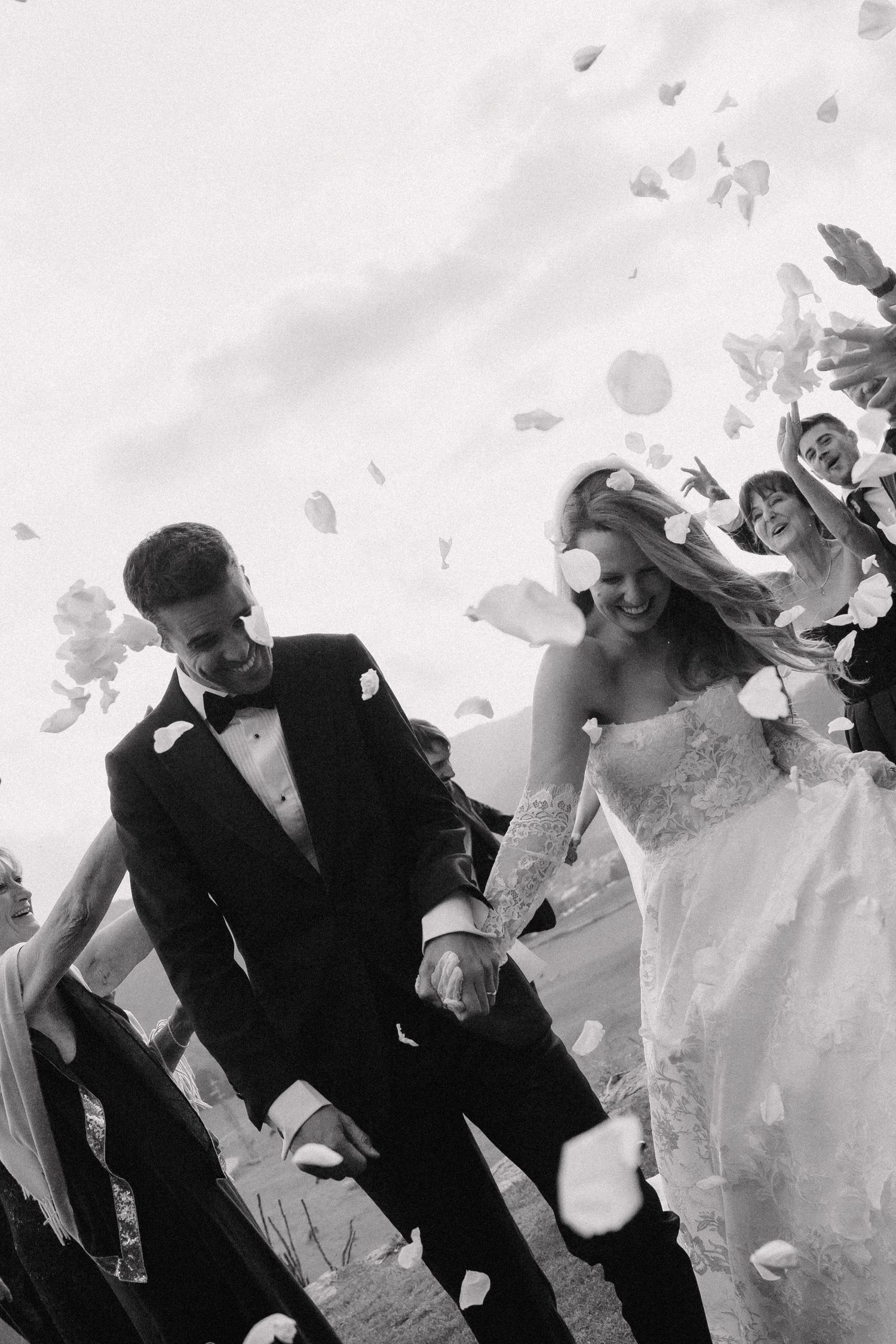 A black and white wedding photo sequence showing a couple joyfully running through falling flower petals outdoors.