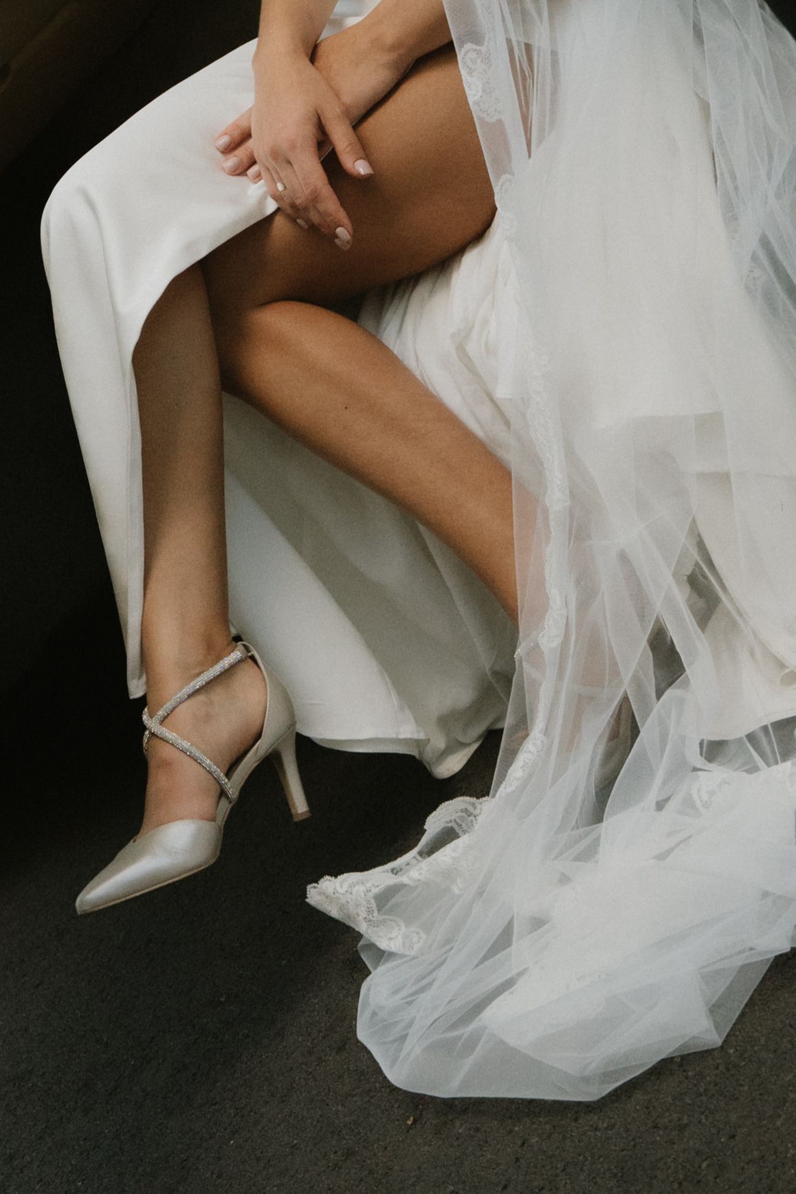 Close-up details of a bride's silver heels and flowing white tulle dress against a dark floor.