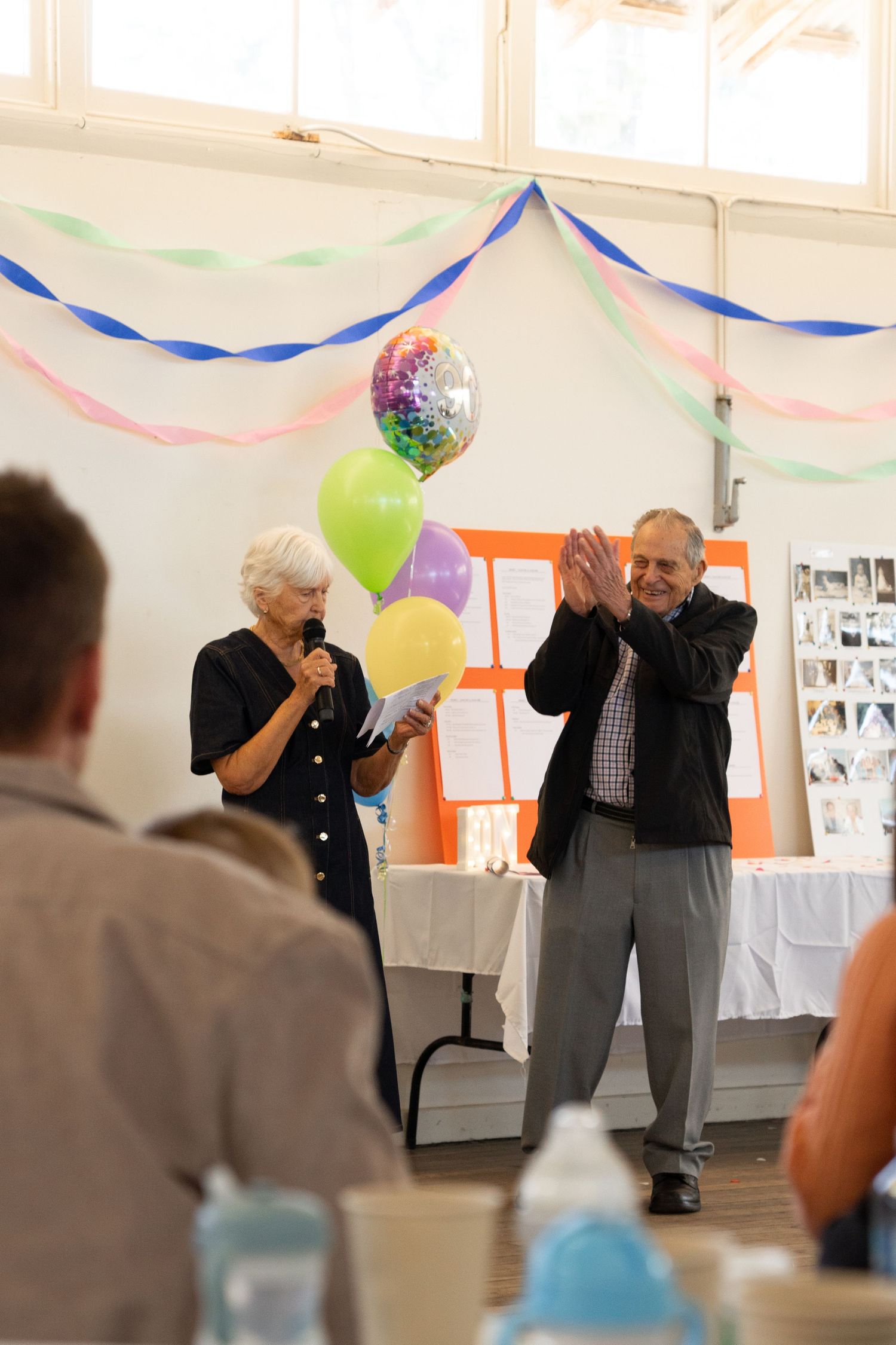Elderly couple celebrating at a party with colorful balloons and streamers, man clapping while woman speaks into microphone.