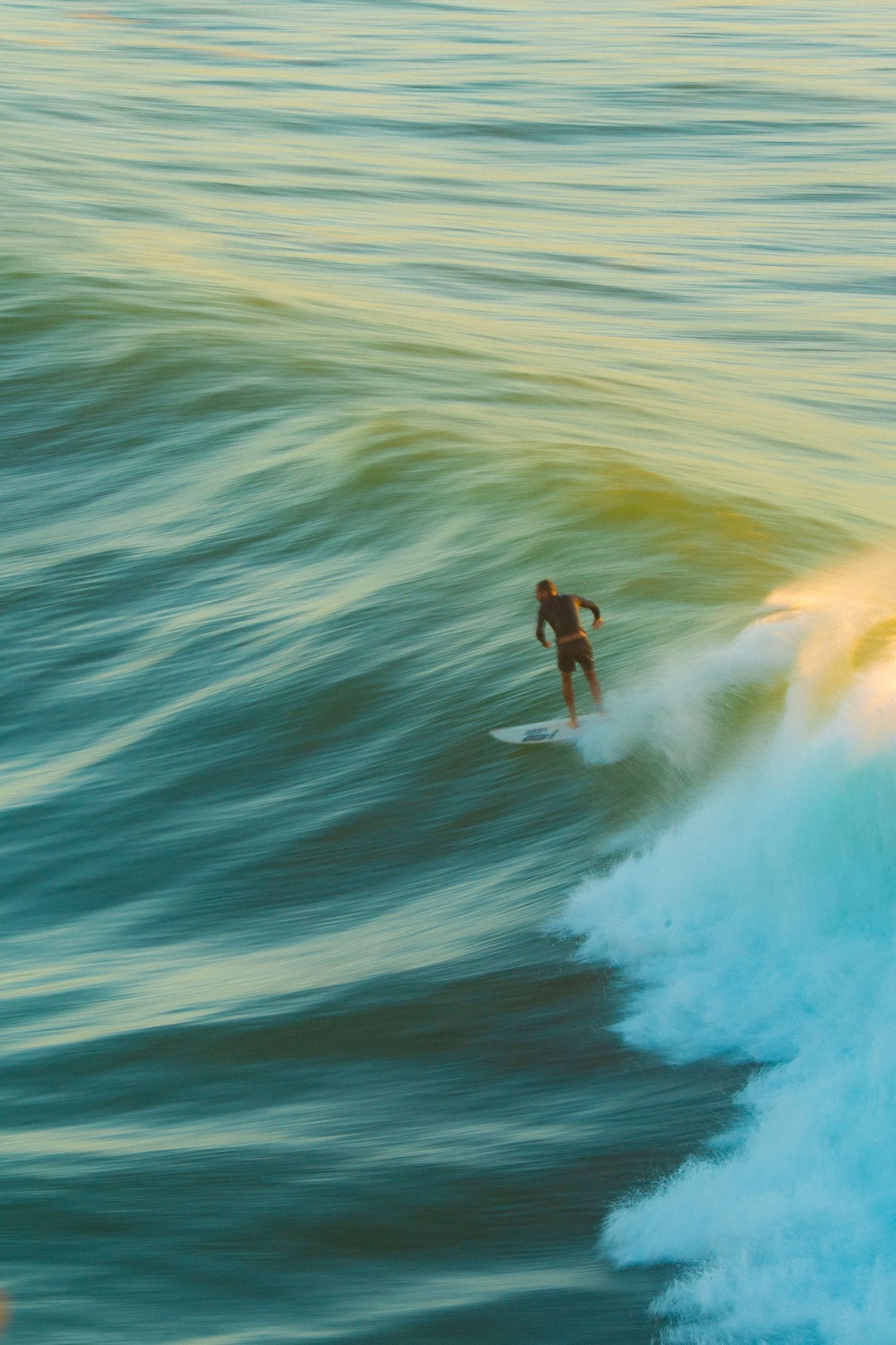 A surfer rides a rolling wave in golden-green ocean waters, captured in a stunning aerial perspective.