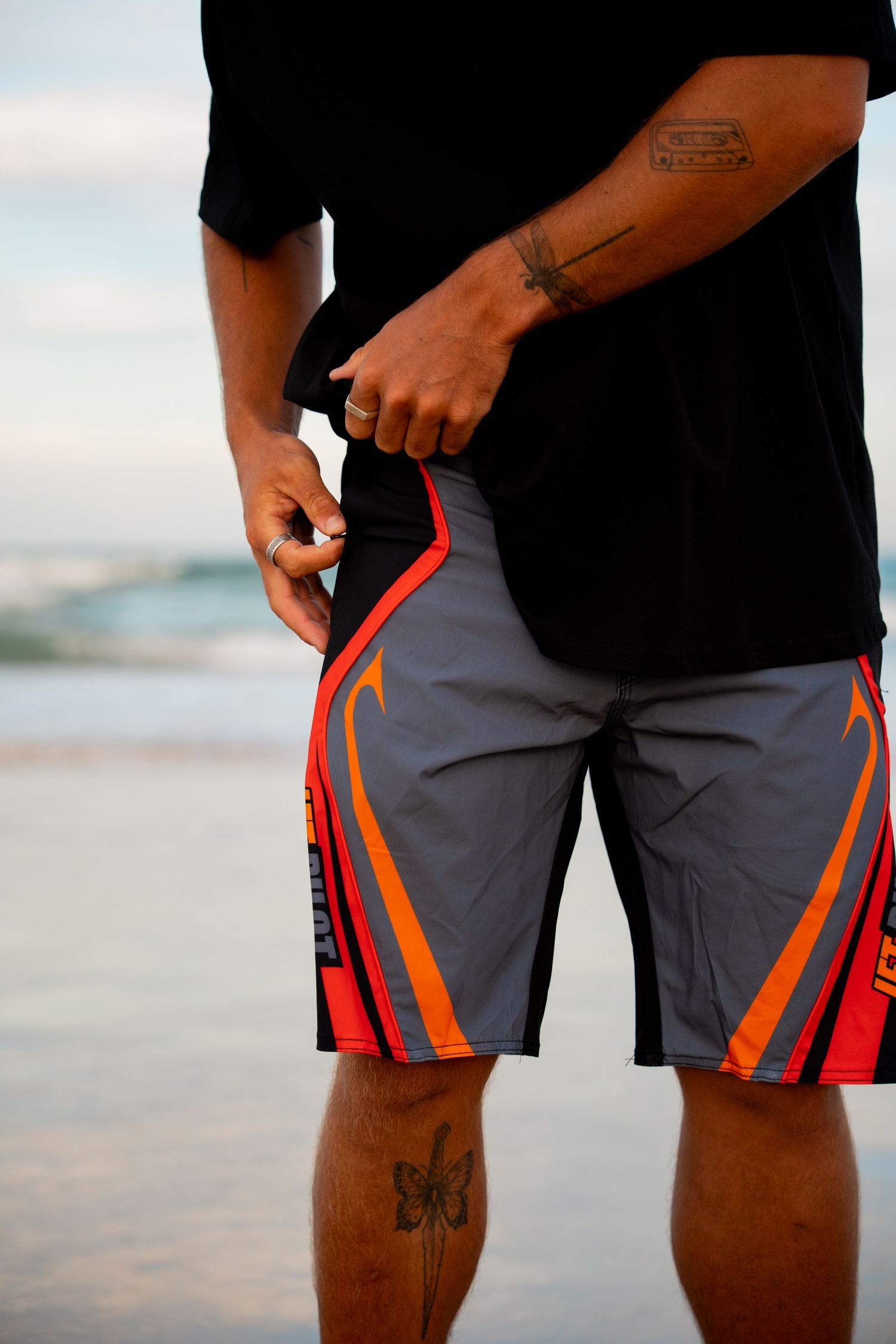 Man wearing gray and red board shorts with orange accents standing on a beach with ocean waves in the background.
