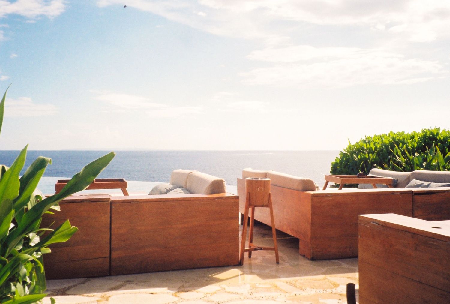 Wooden outdoor lounge furniture on a terrace overlooking a calm blue ocean under a clear sky.