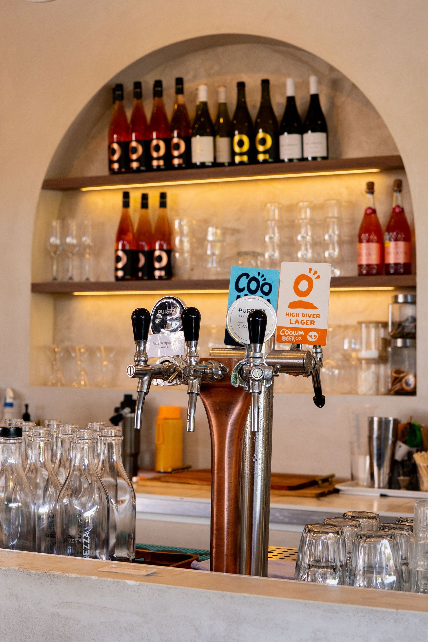 Beer taps and glassware at a bar with illuminated shelves displaying wine bottles and spirits in an arched alcove.