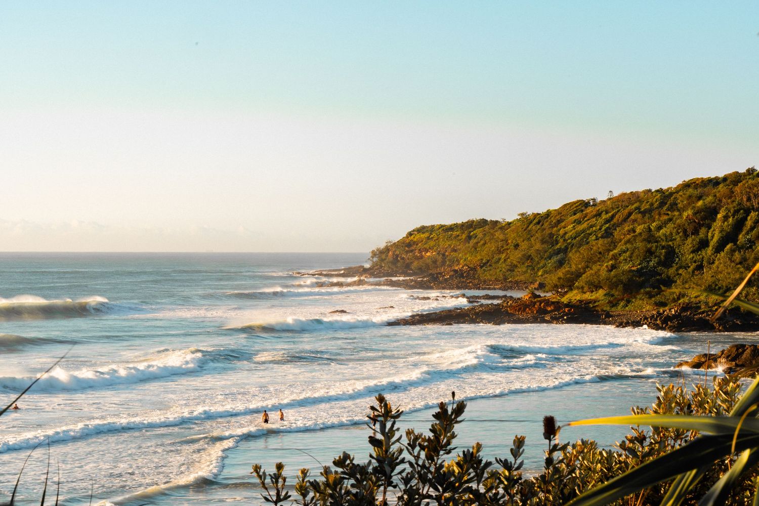 Scenic coastal view with rolling ocean waves, lush green headland, and a sandy beach under a clear blue sky.