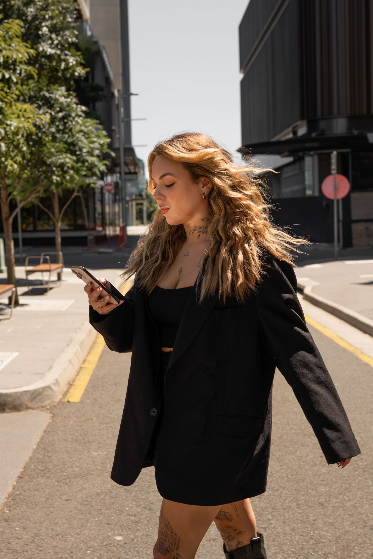 Young blonde woman with tattoos in black blazer and skirt walks urban street while looking at smartphone.
