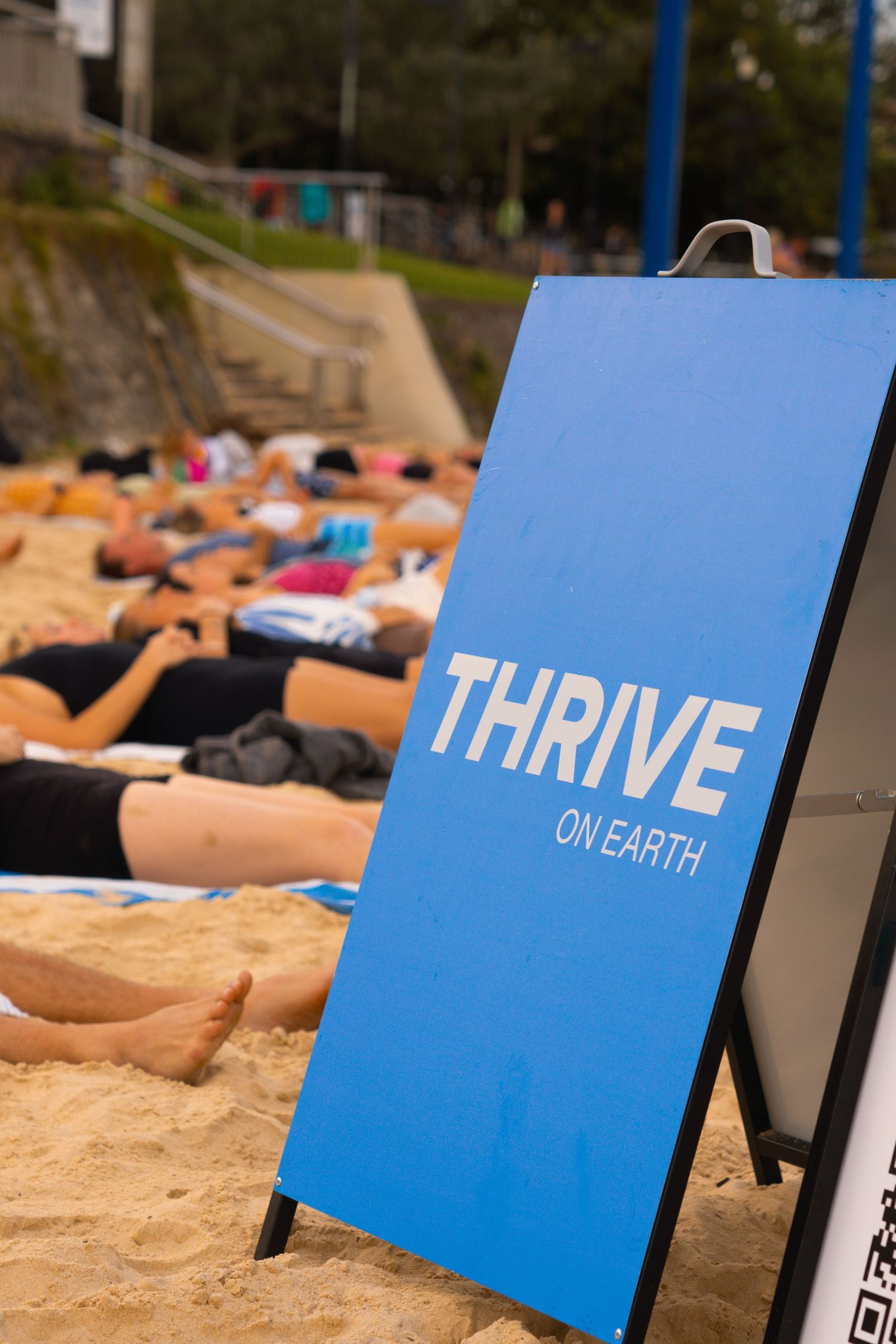 Blue Thrive on Earth sign on sandy beach with people sunbathing in the background.