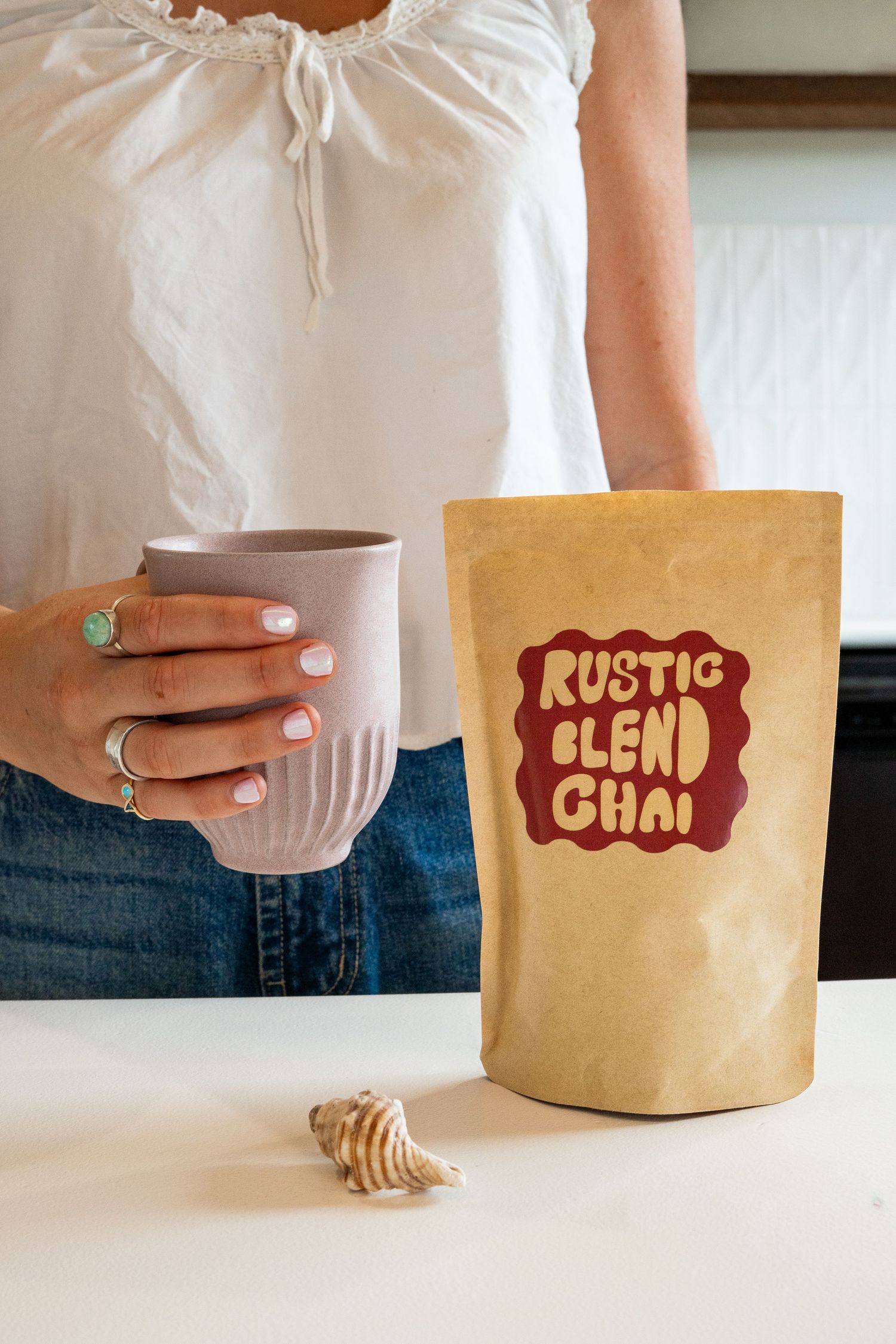 Woman holding a pink mug next to a kraft paper bag of Rustic Blend Chai tea on a white surface.