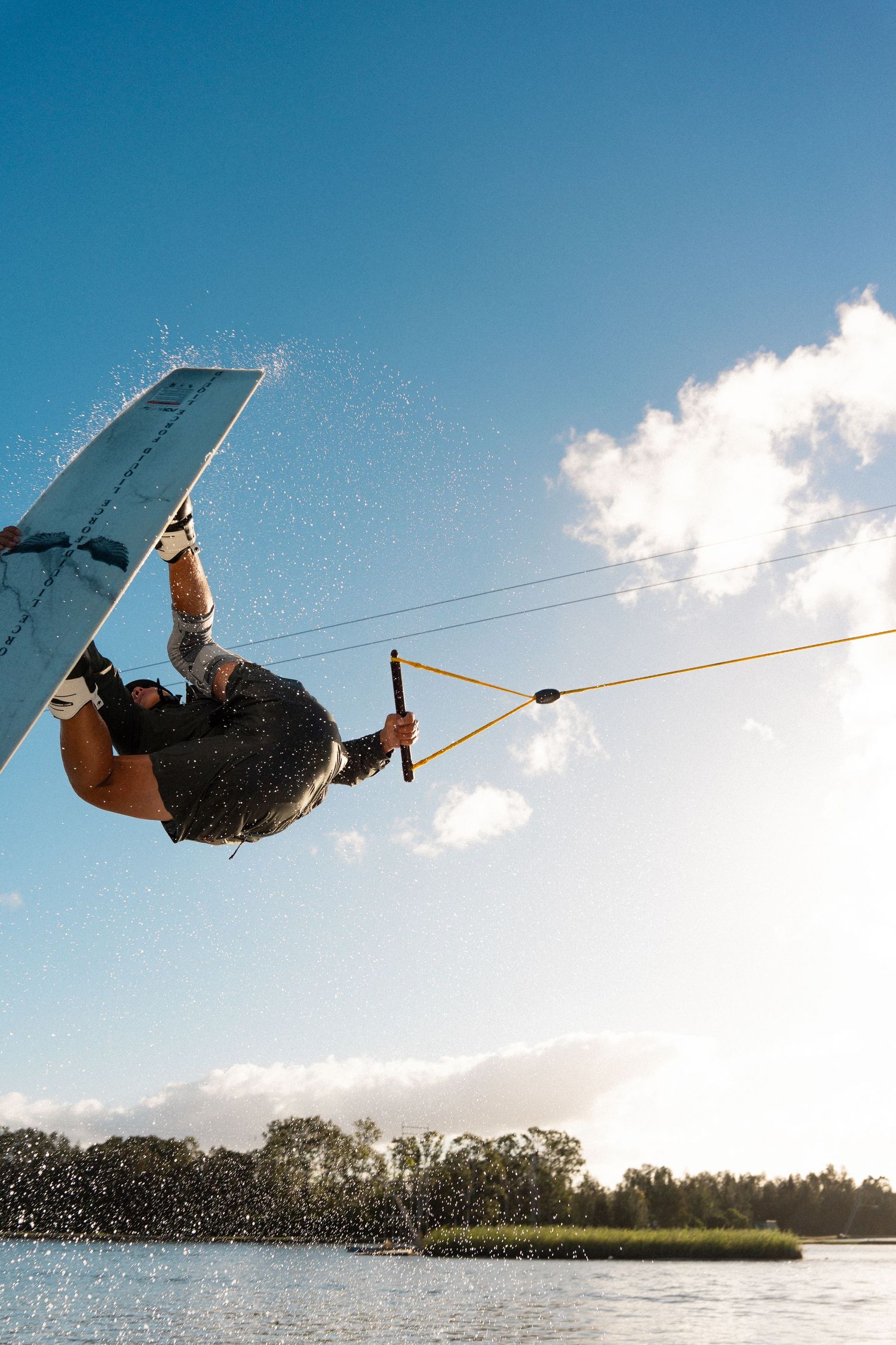 A wakeboarder performs an inverted trick mid-air while holding a tow rope against a bright blue sky over a calm lake.