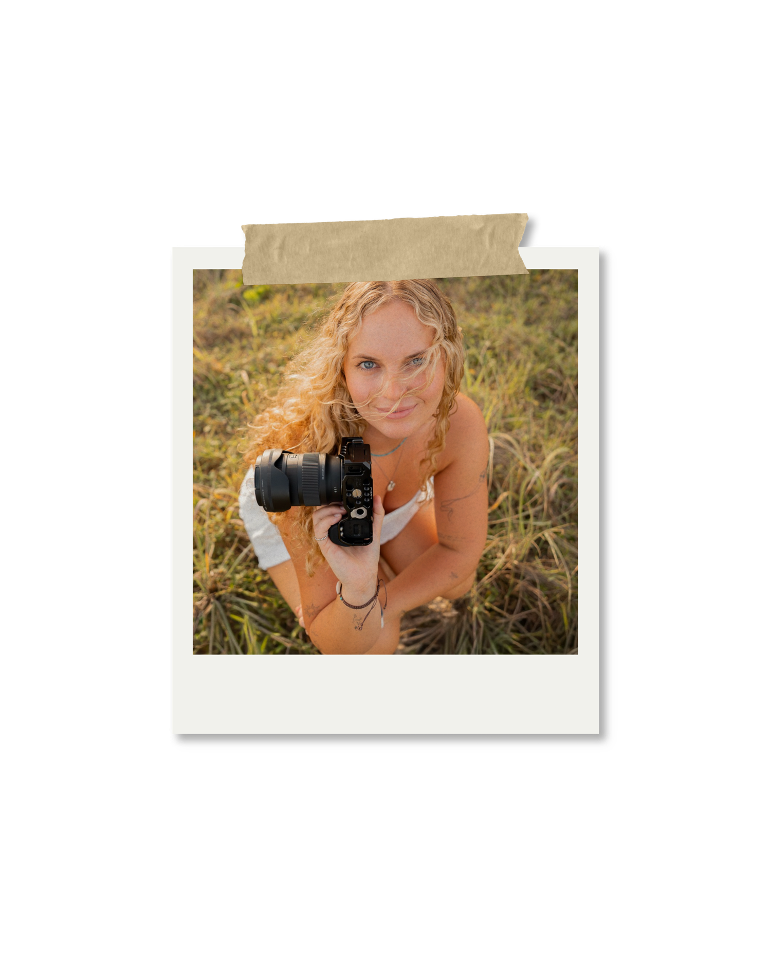 A smiling woman with curly blonde hair holds a DSLR camera outdoors in a grassy field, looking up at the viewer.