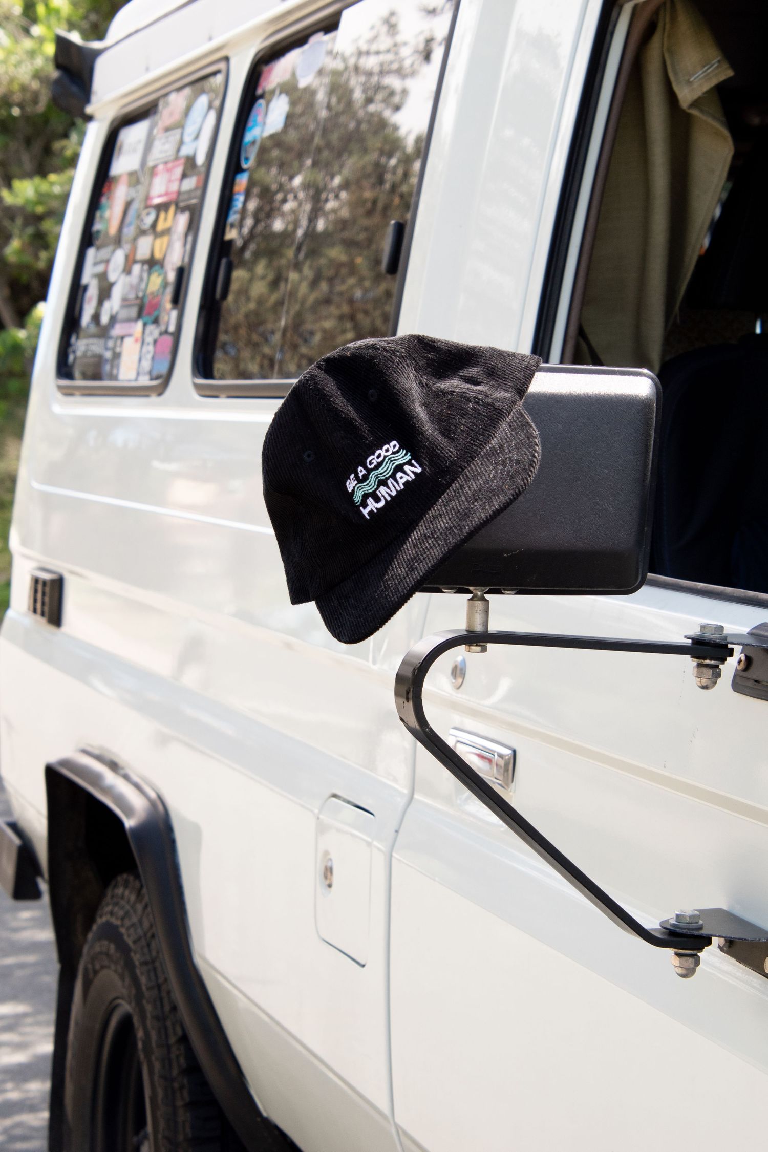 Black beanie hat resting on white van side mirror, with sticker-covered windows visible in background.
