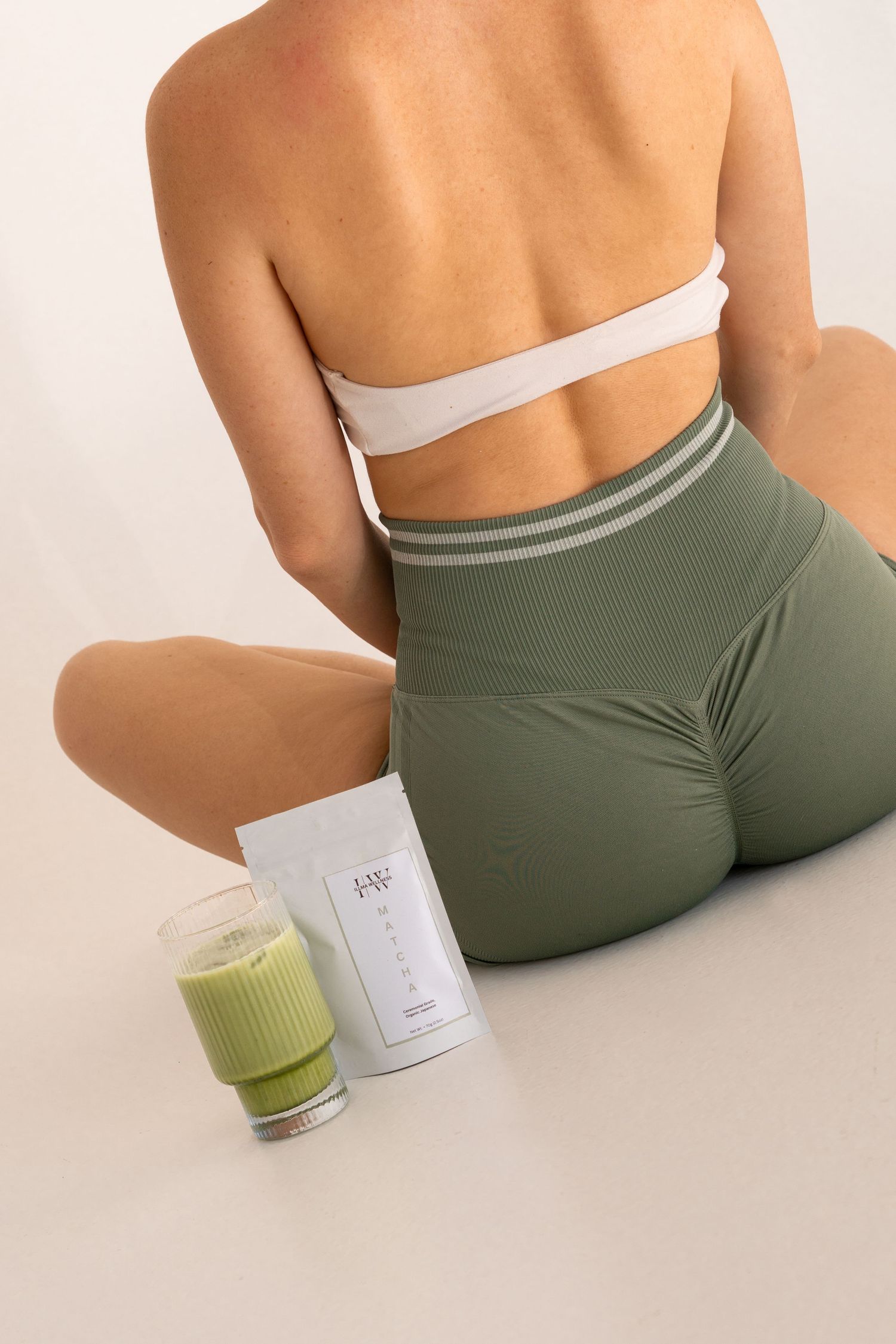 Woman in olive green activewear sitting beside a green smoothie and white supplement powder pouch.