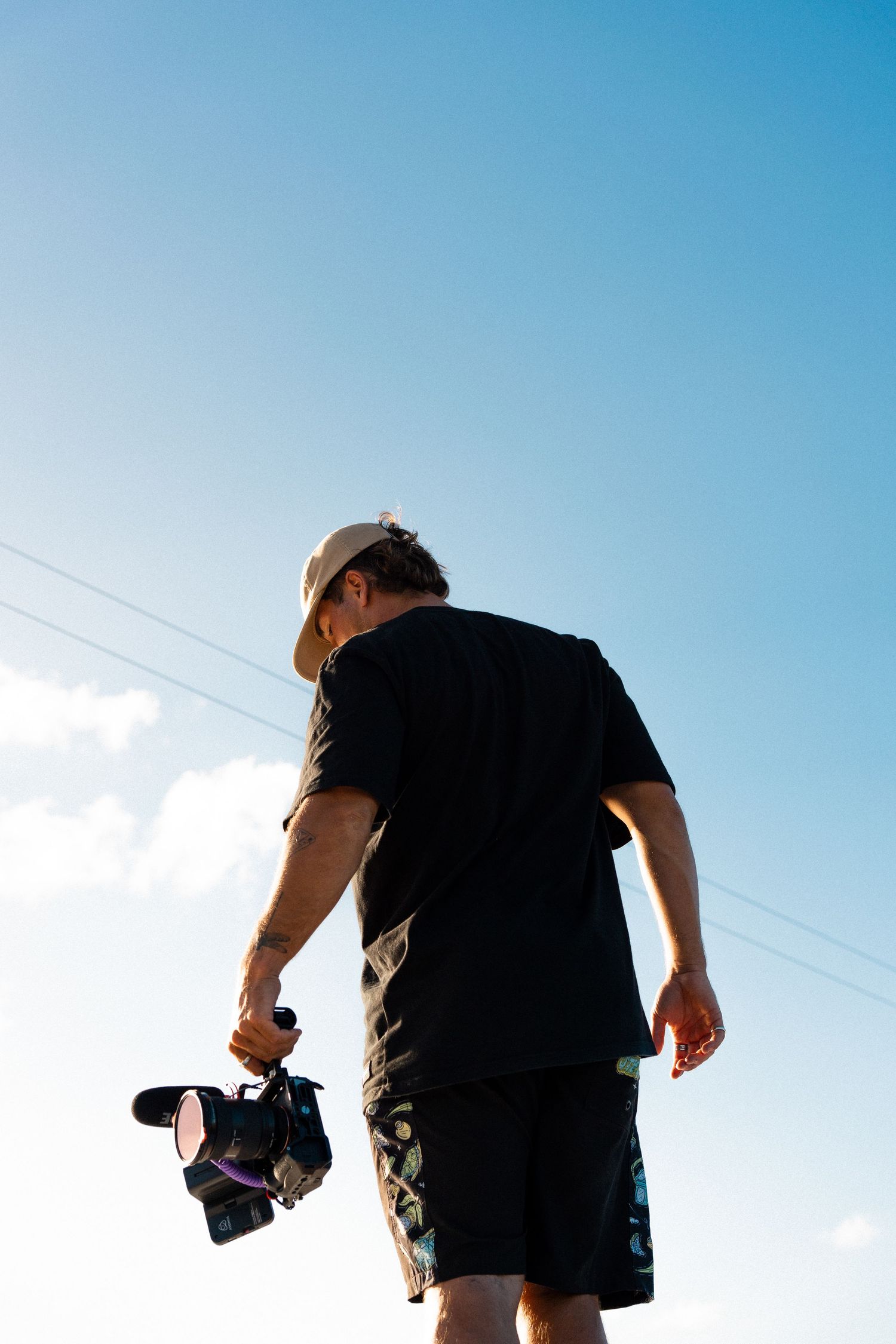 Man in black t-shirt and cap holds camera, shot from below against blue sky with power lines.