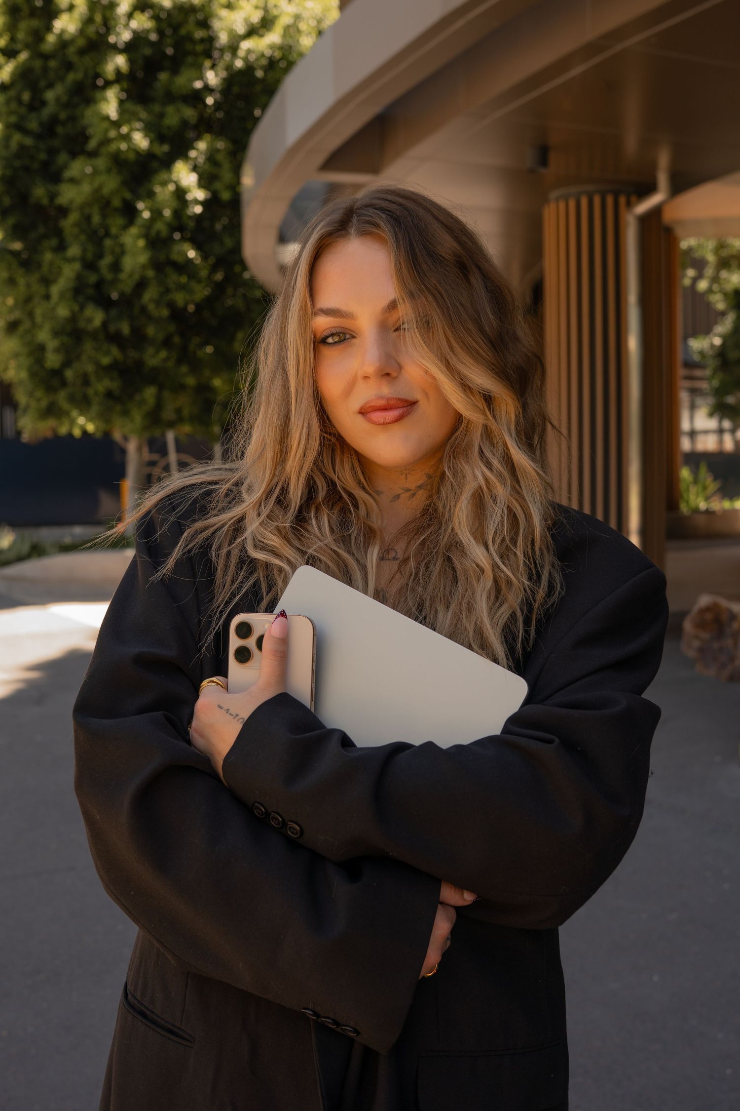 Young blonde woman in black coat holding laptop and phone outdoors in warm sunlight near modern building.