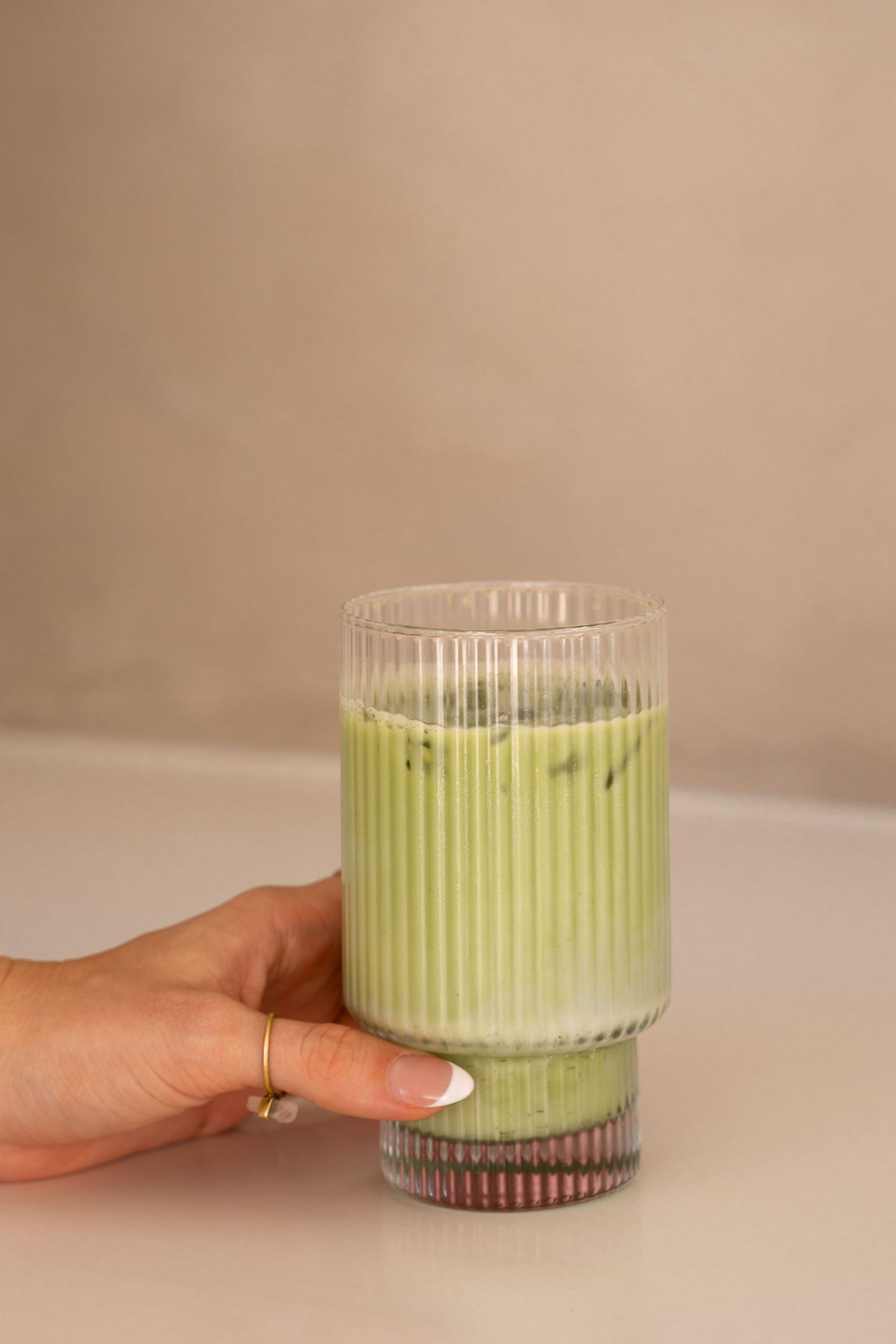 Hand holding a ribbed glass with iced matcha latte on a neutral background.