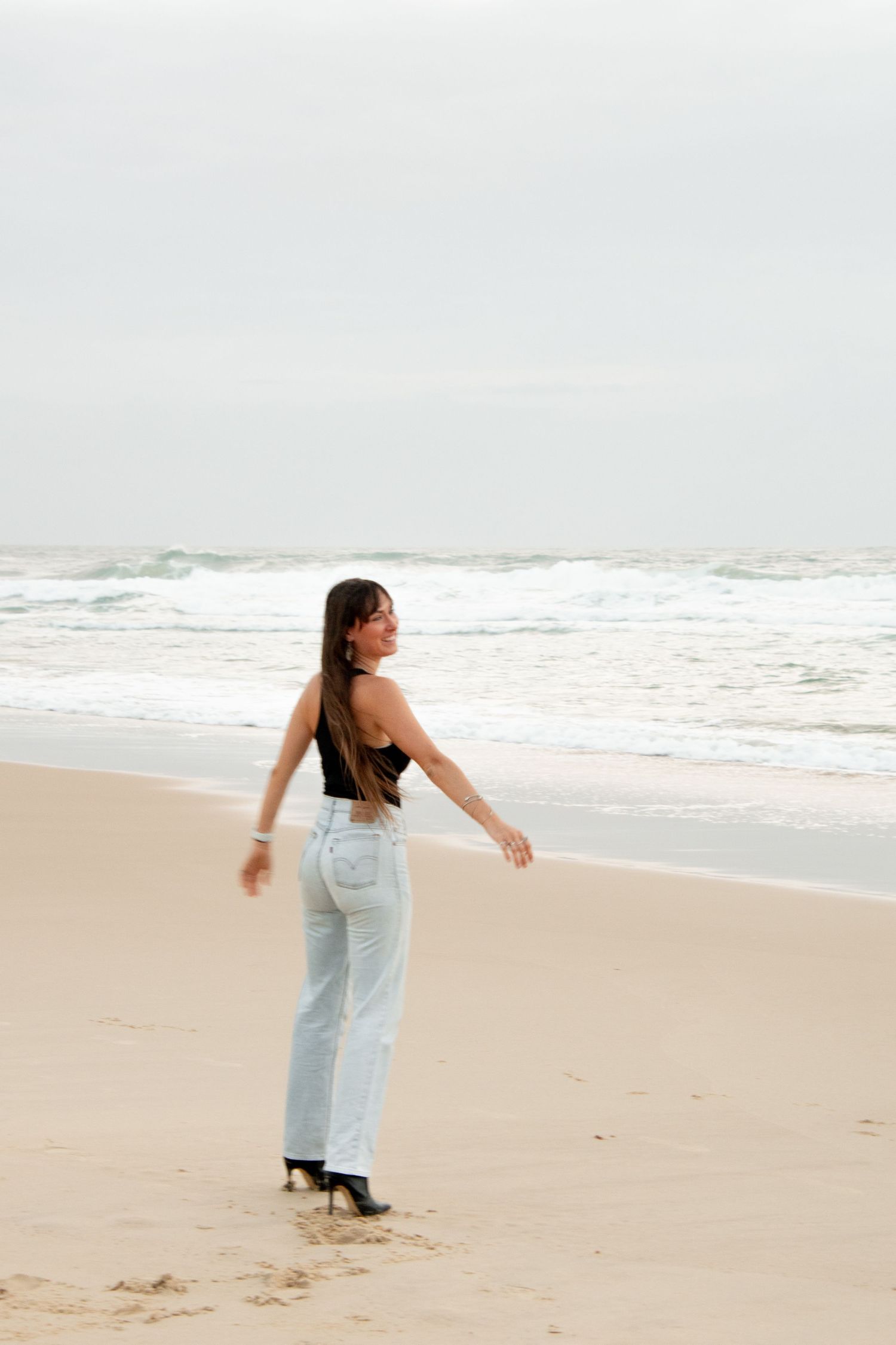 Woman in black crop top and light blue wide-leg jeans standing on sandy beach with ocean waves in background.