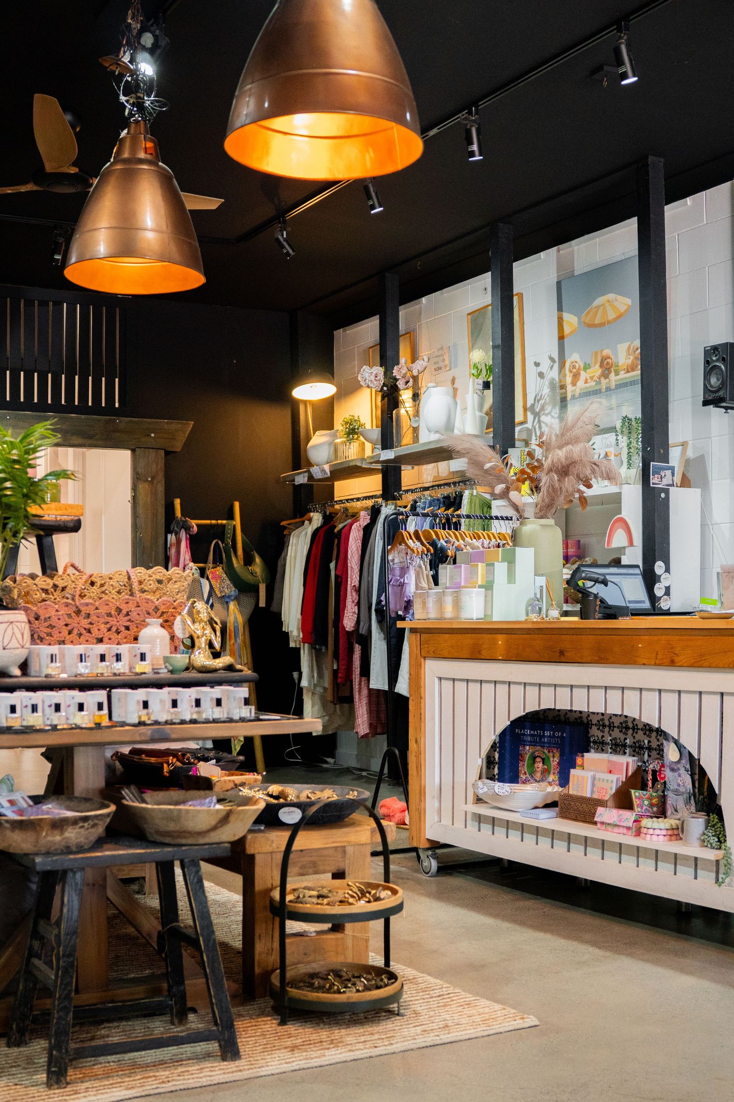 Interior of a boutique retail store with copper pendant lights, clothing racks, and rustic display tables with merchandise.
