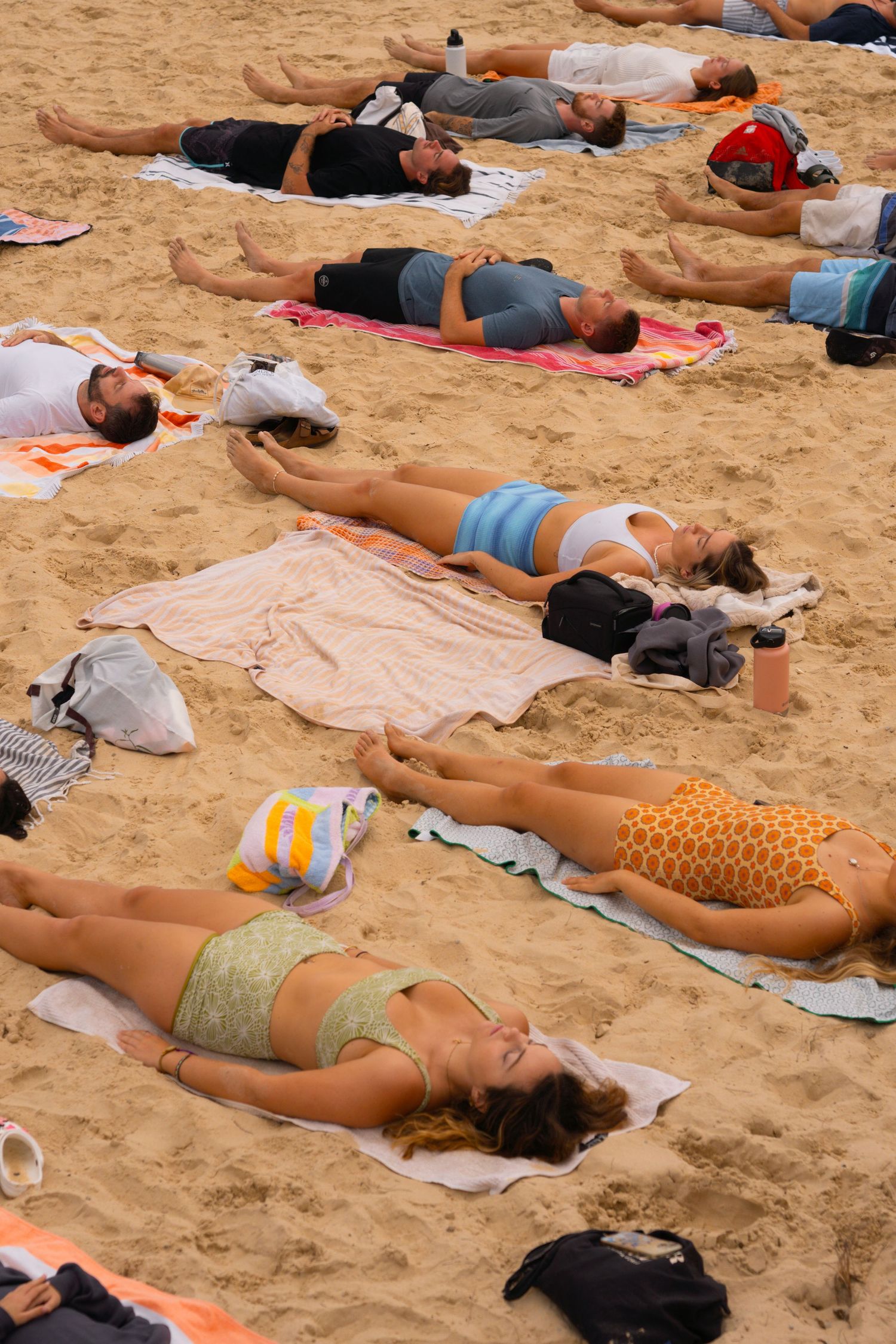 Group of people lying on towels on sandy beach doing yoga savasana pose outdoors.